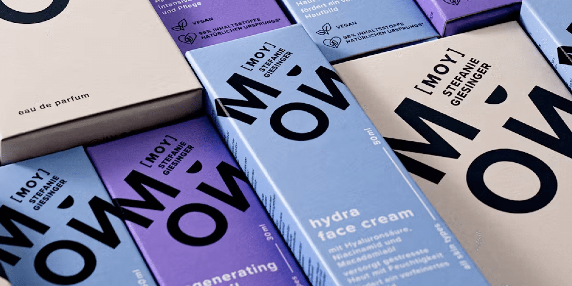 Markenwelt MOY by Stefanie Gieringer – vegane Skincare- und Parfum-Verpackungen in Lavendel, Flieder und Beige mit grosser typografischer „MOY"-Wortmarke, diagonal arrangiert.