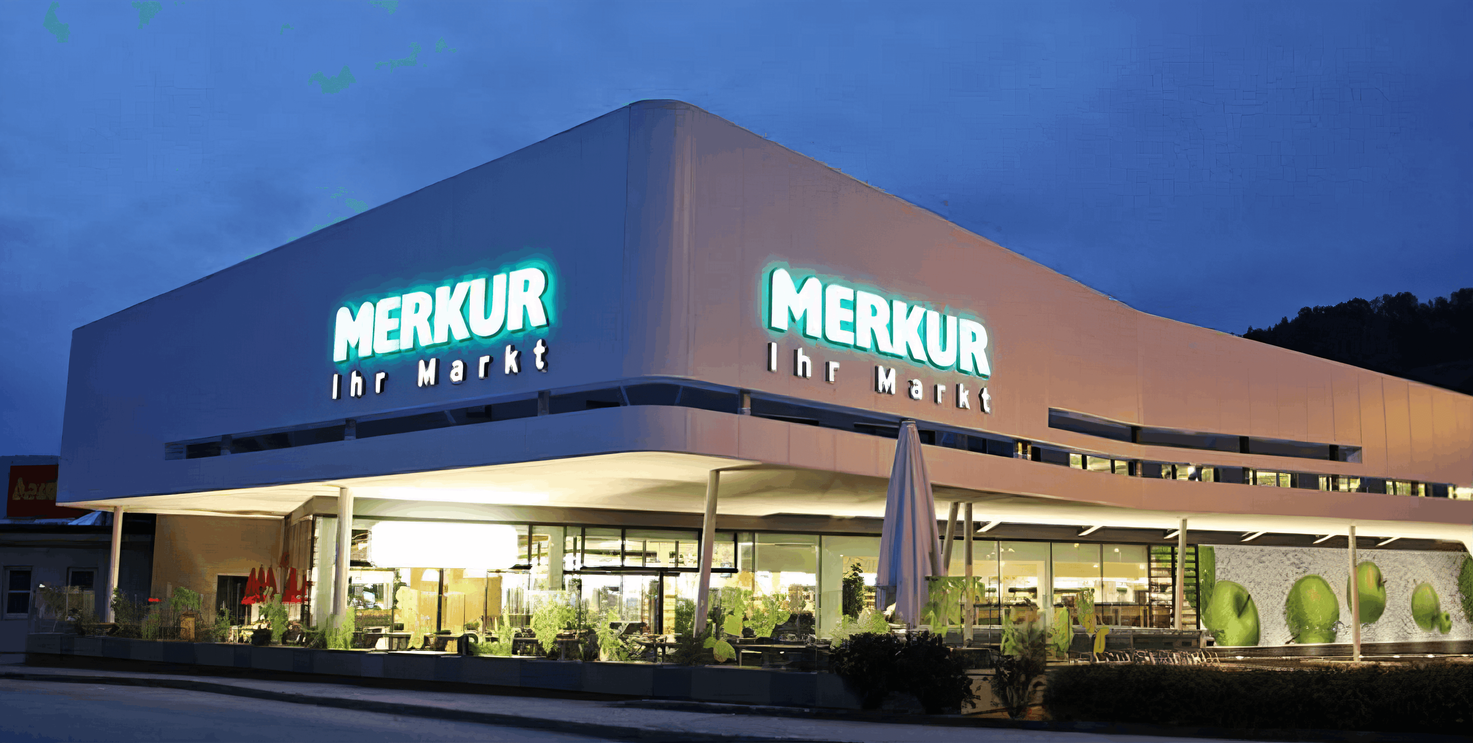 MW Interior - Merkur Ihr Markt Supermarkt bei Nacht mit beleuchteter türkiser Außenbeschilderung und Glasfront" Beschreibung: Merkur Supermarkt-Standort in Abendbeleuchtung mit charakteristischer türkiser Außenbeschilderung und großzügiger Verglasung – ein Referenzkunde, für dessen Filialkonzepte Einrichtungs- und Möbellösungen umgesetzt wurden.
