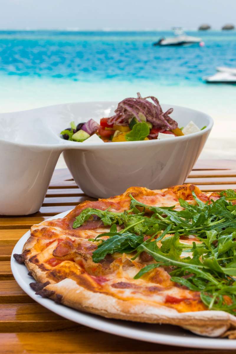A pizza topped with arugula and tomatoes sits on a wooden table in front of a bowl of salad by a tropical beach.