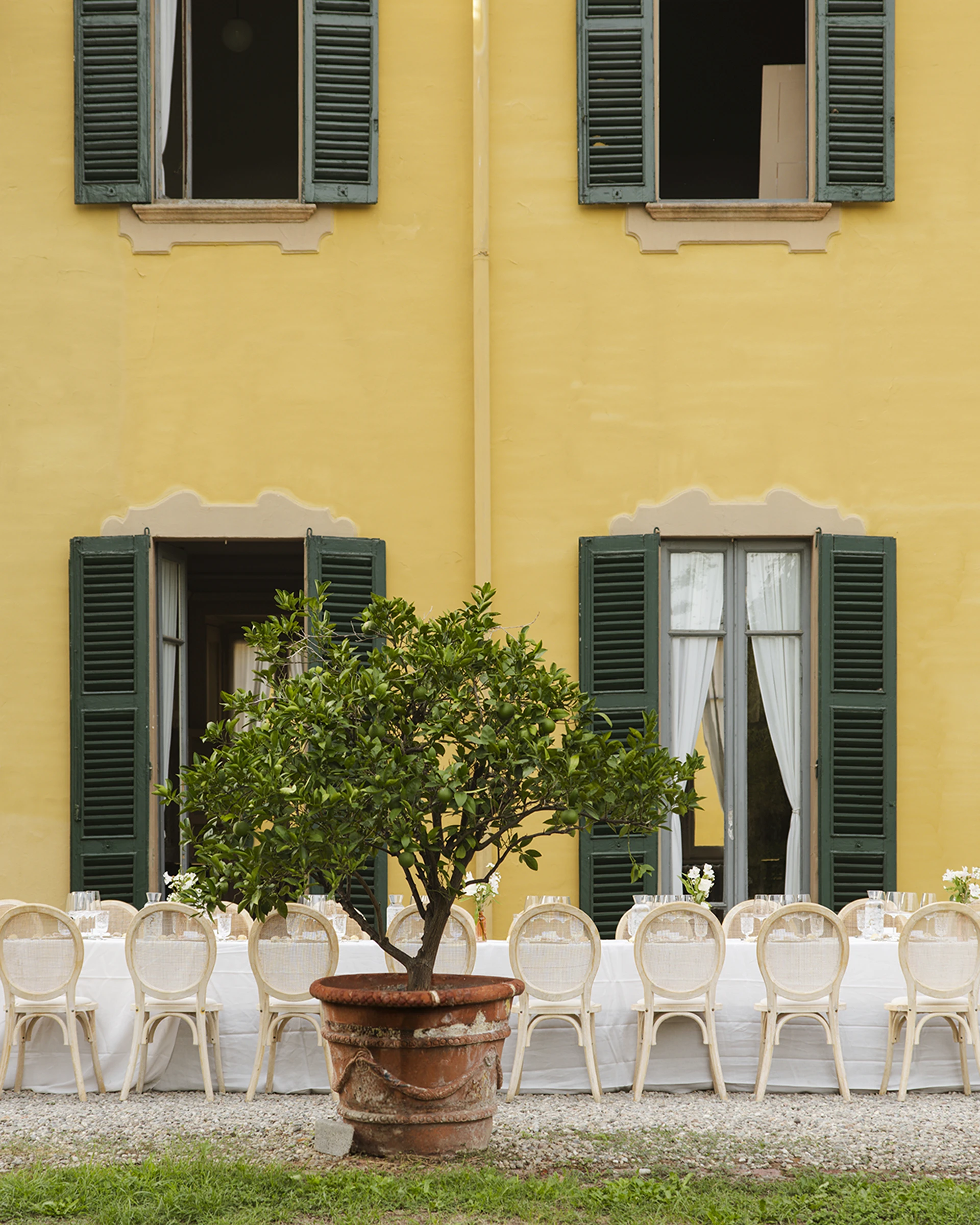 Facciata gialla con persiane verdi, limone in vaso di terracotta e tavolata apparecchiata con sedie chiare