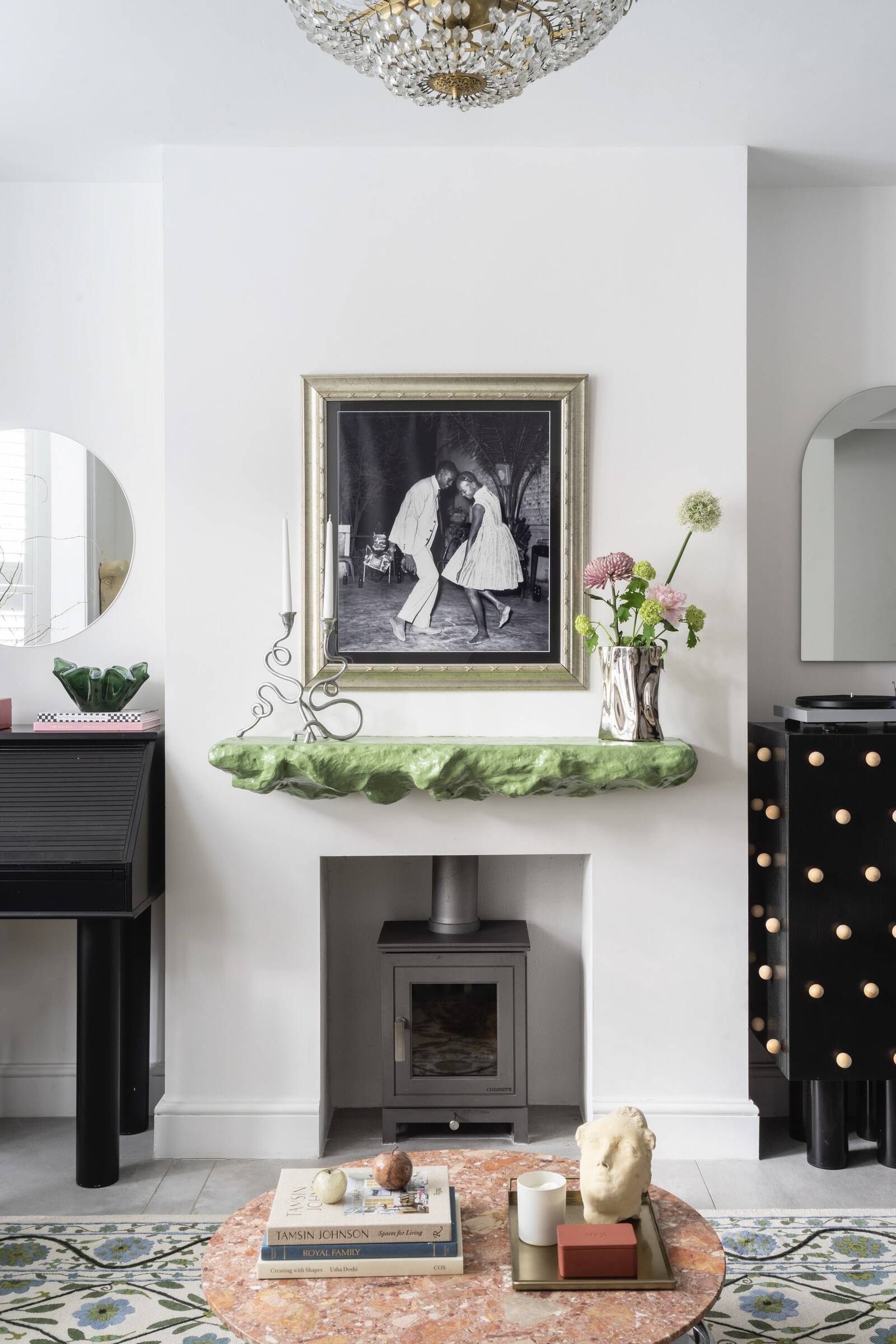 A fireplace with a green sculptural mantlepiece, holding a chrome vase, flowers and unique candlestick holders. Above there is artwork of people dancing.