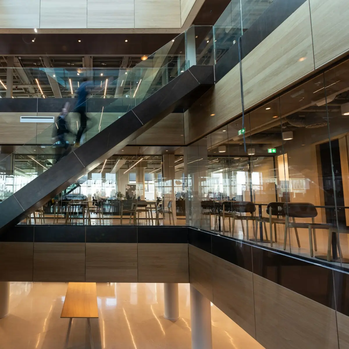 Modern office interior with glass walls, wooden accents, and a person walking up a staircase in motion blur.