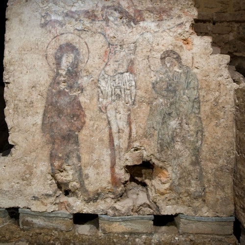 Ancient mural of three faded figures, including a central crucified figure, on a rough stone wall, partially deteriorated.