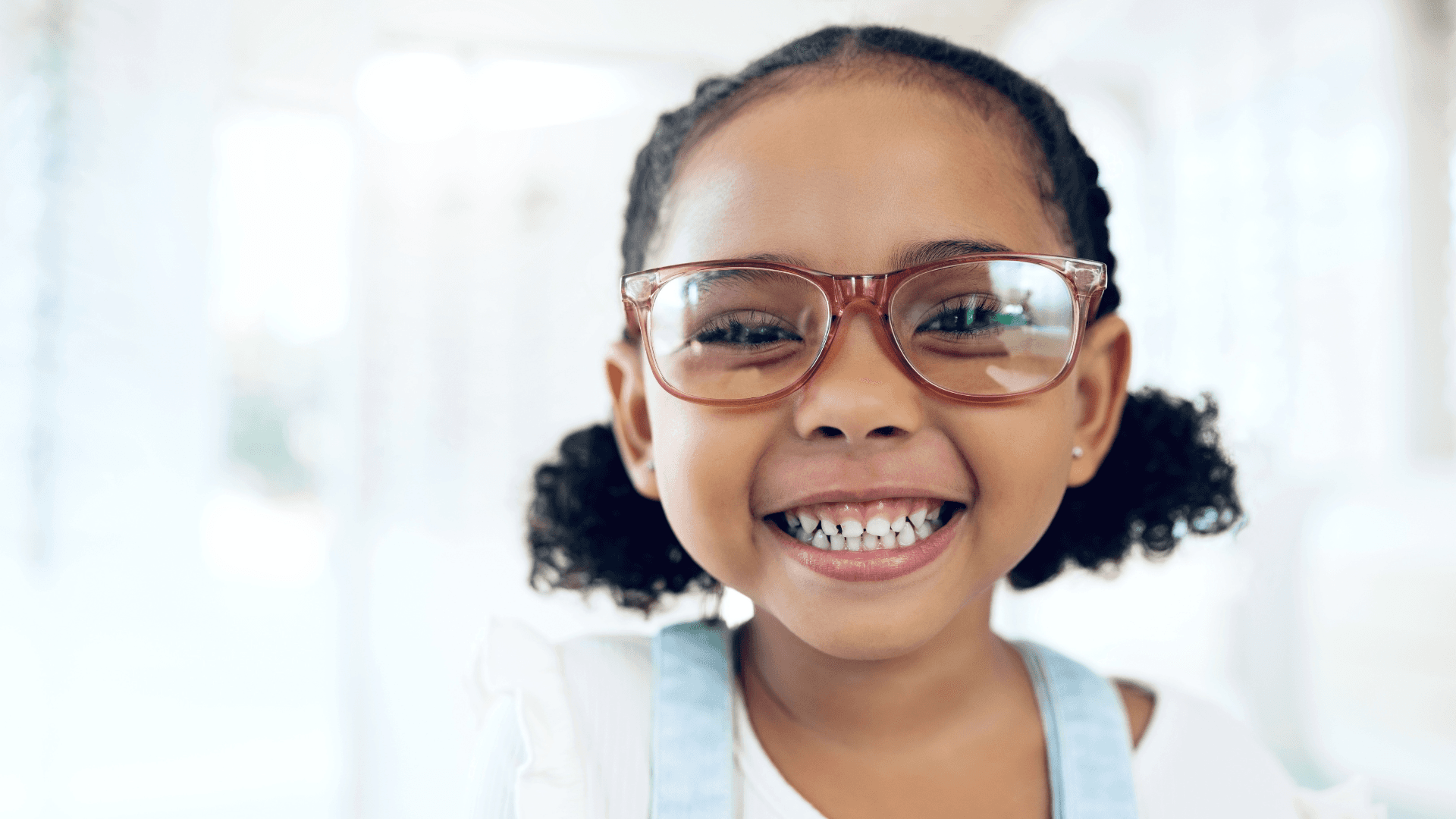 a young child with pigtails smiles wide into the camera with reddish brown rimmed glasses and a blurred out white background
