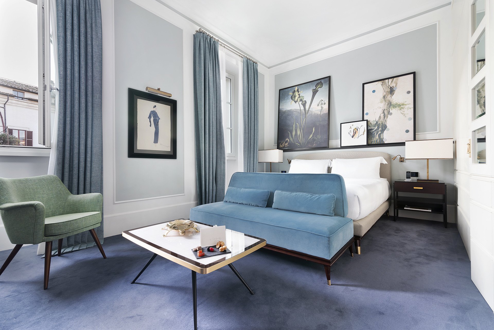 A modern hotel room with blue walls and a blue sofa, featuring framed art, a white bed, and a small dining table.