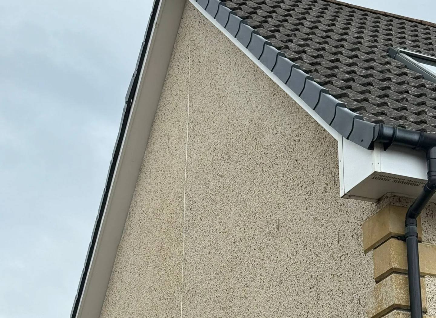 A beige house wall with textured stucco, featuring a sloped roof with dark, wavy tiles. A black gutter runs down, against a cloudy sky.