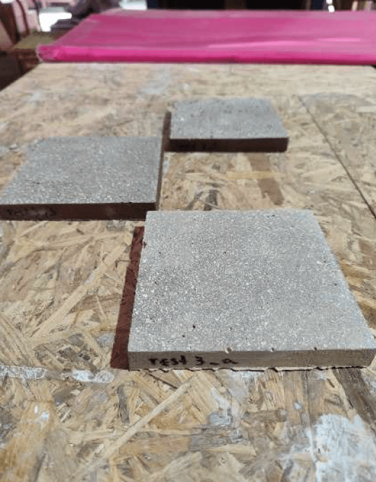 Deux dalles carrées en matériau composite gris granuleux étiquetées, posées sur une planche en OSB, montrant une surface fine sans éclats apparents