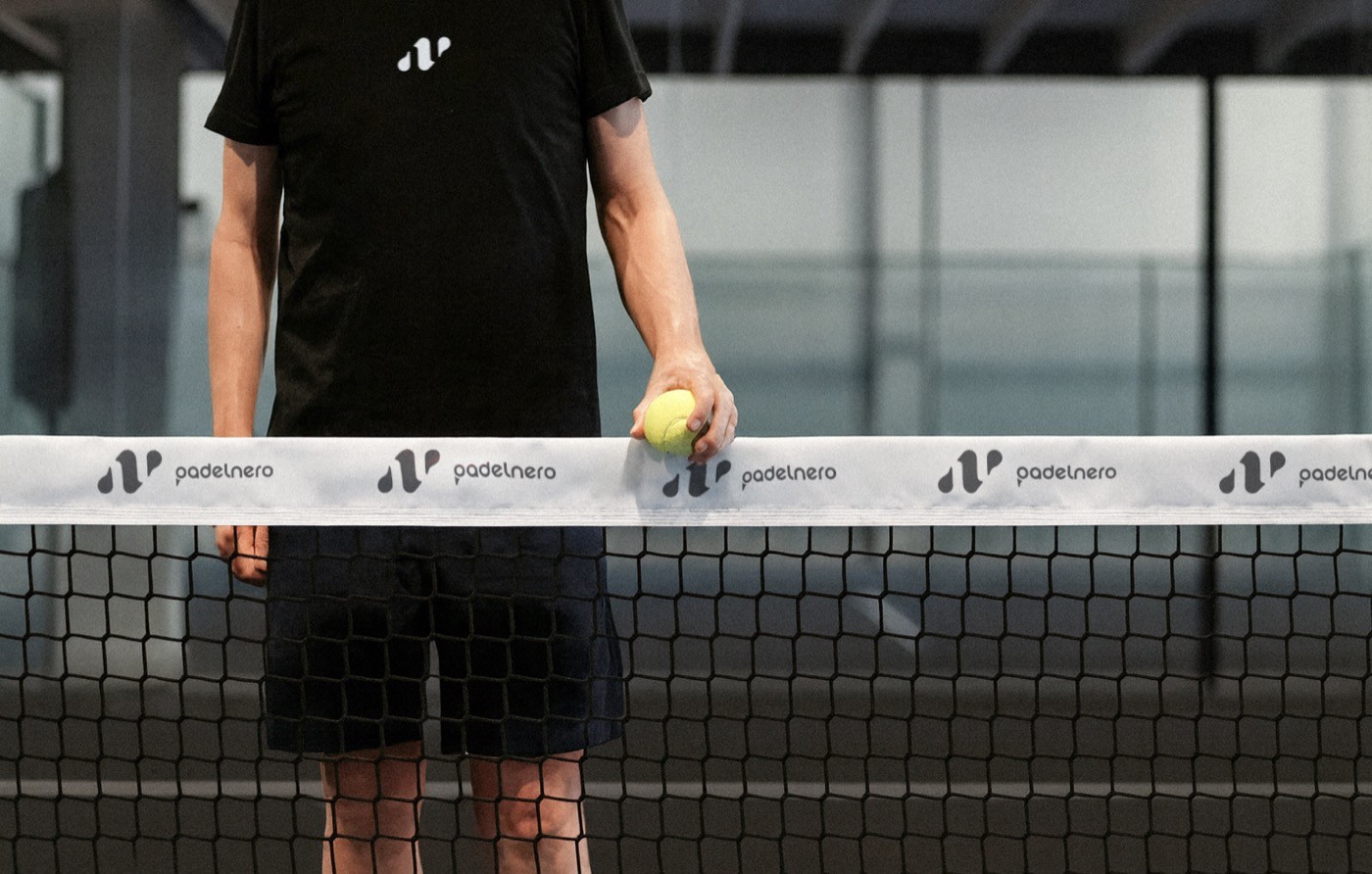Padelspieler steht am Netz mit einem Ball in der Hand, auf der Netzbande ist das Padelnero-Logo zu sehen.