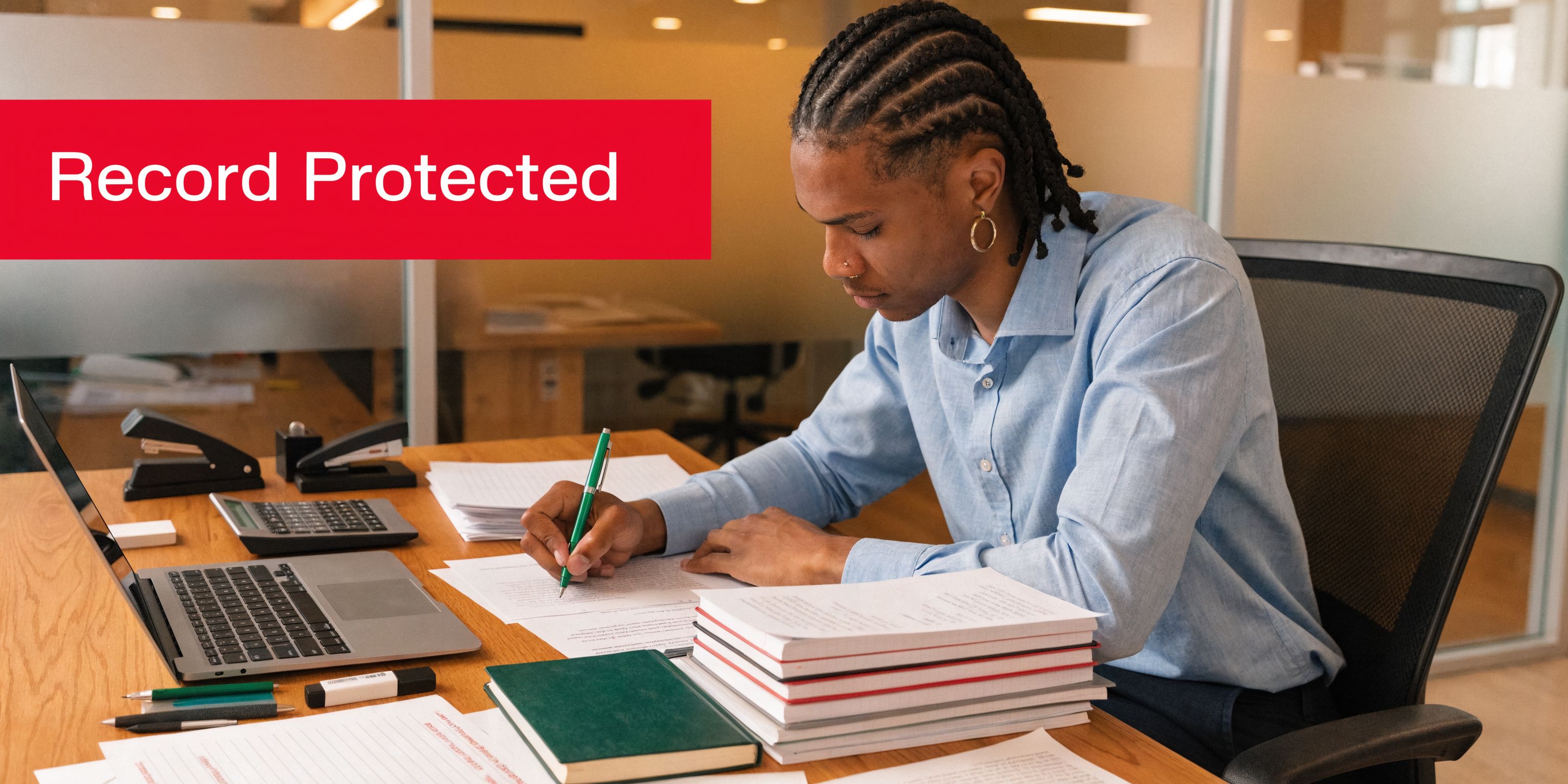 A focused professional with braided hair writing on documents at an office desk with a laptop.