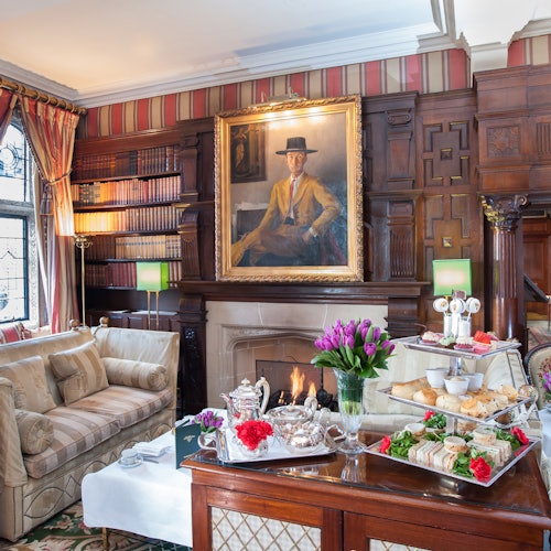 Elegantes Zimmer mit einem Kamin, einem Gemälde darüber, Bücherregalen, gemütlichen Sitzgelegenheiten und einem mit Nachmittagstee und Blumen gedeckten Tisch.