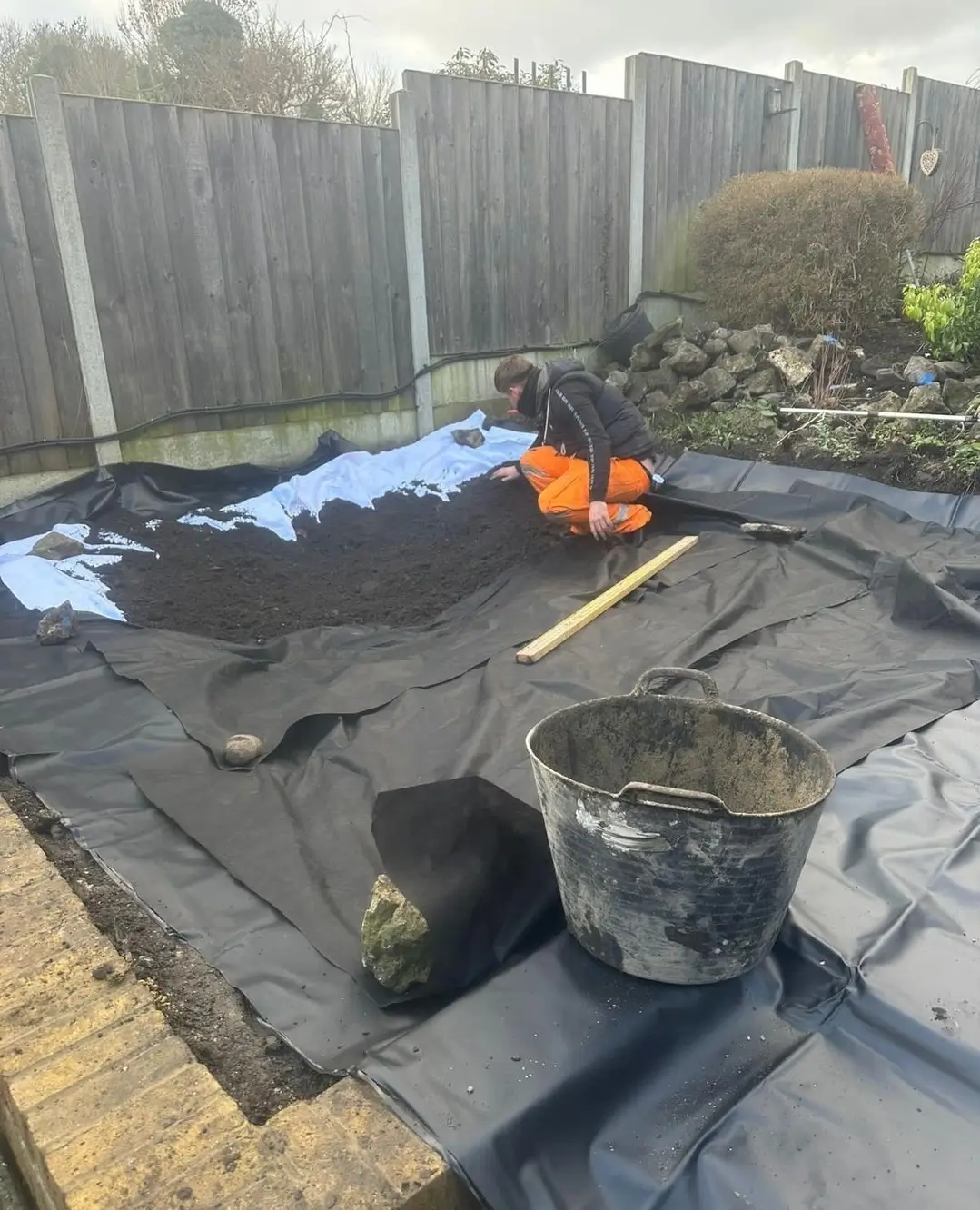 A garden area under construction, featuring black landscaping fabric, stones, and small piles of soil.