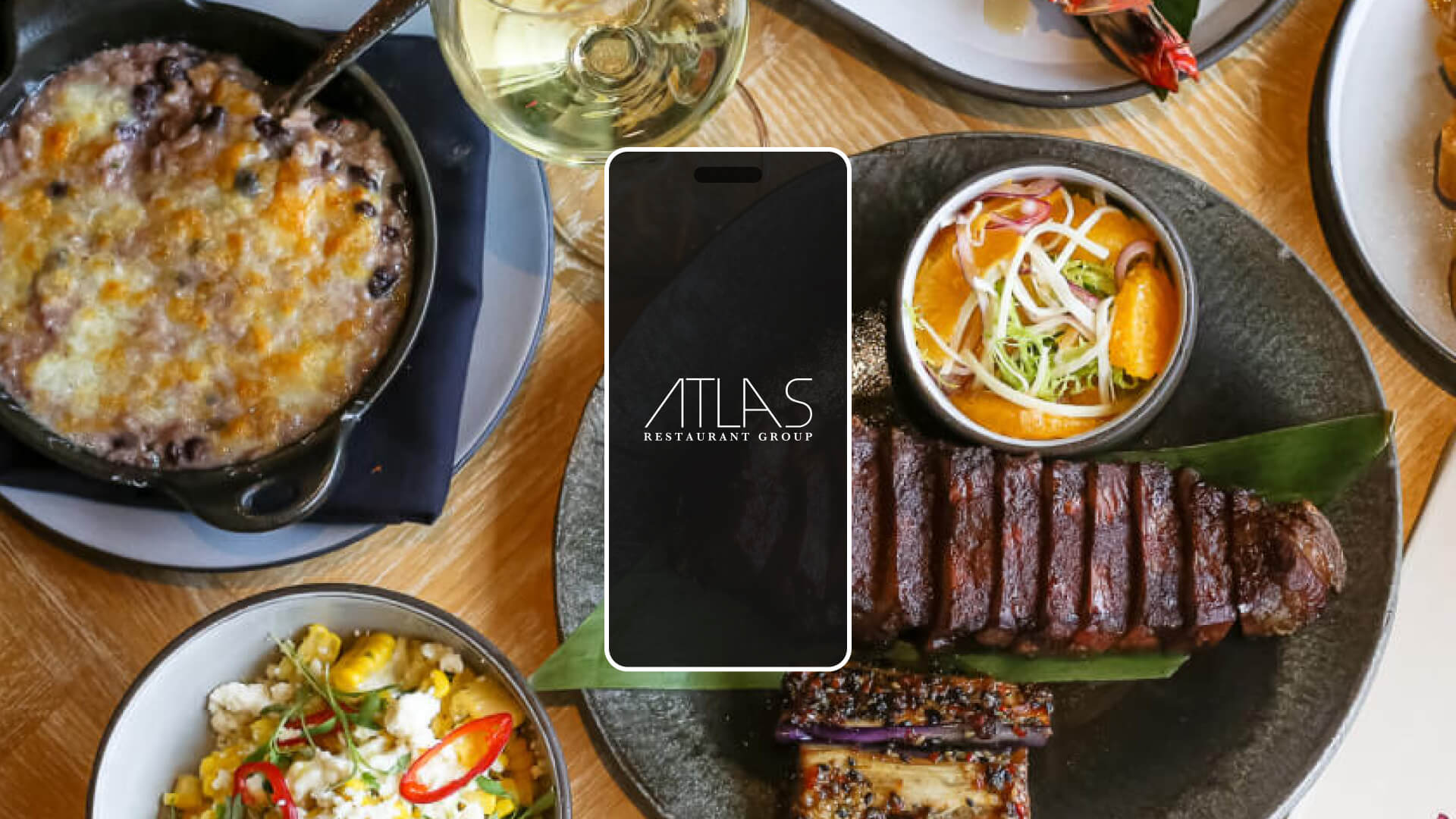 A table set with various plated dishes, including grilled steak, casserole, vegetables, a glass of white wine, and a phone displaying the Atlas Restaurant Group logo.
