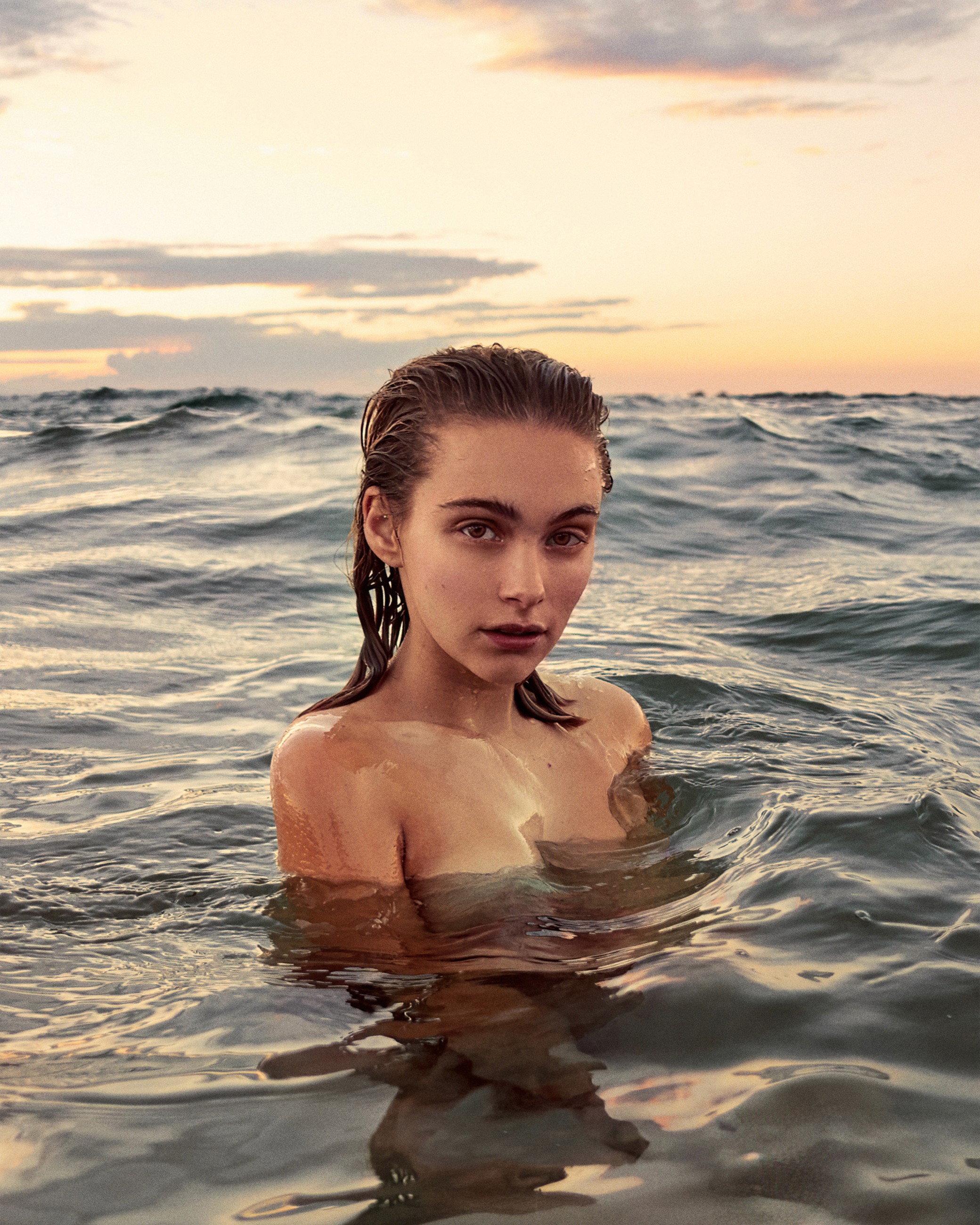 Woman chest-deep in ocean water at sunset with wet hair and bare shoulders, waves and dramatic sky in background.