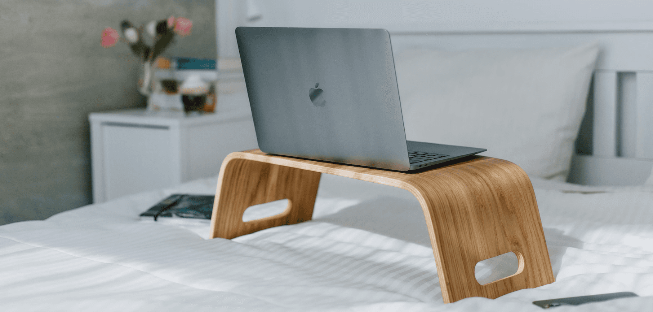A sleek wooden laptop stand on a neatly made bed, holding a space gray MacBook with a cozy, minimalist bedroom setup in the background.