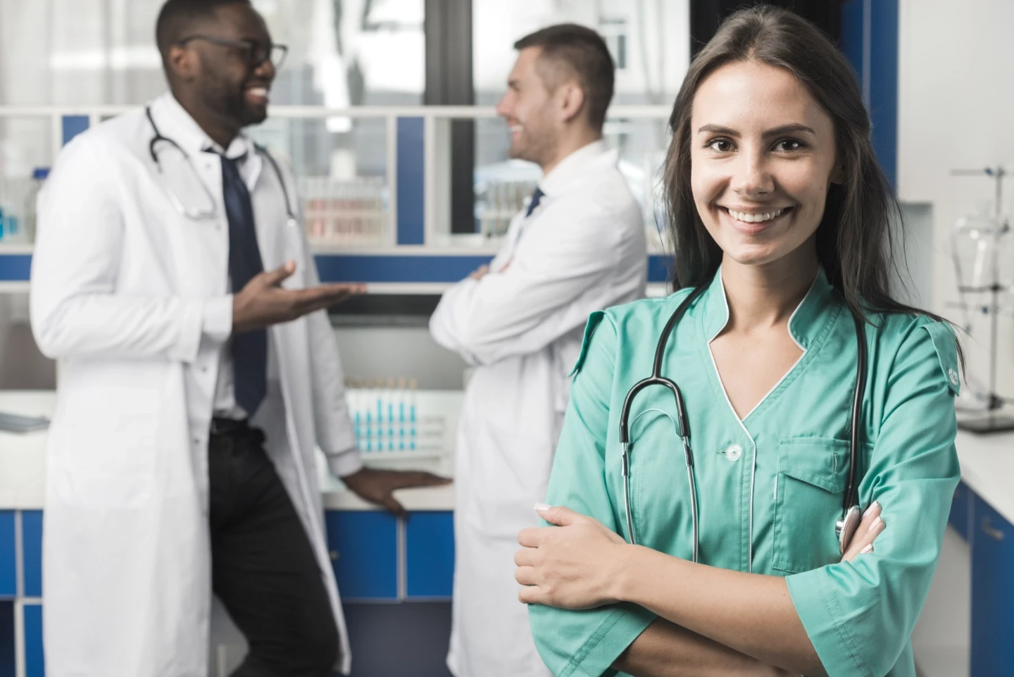 Smiling female resident wearing teal scrubs with two male resident conversing in the background.