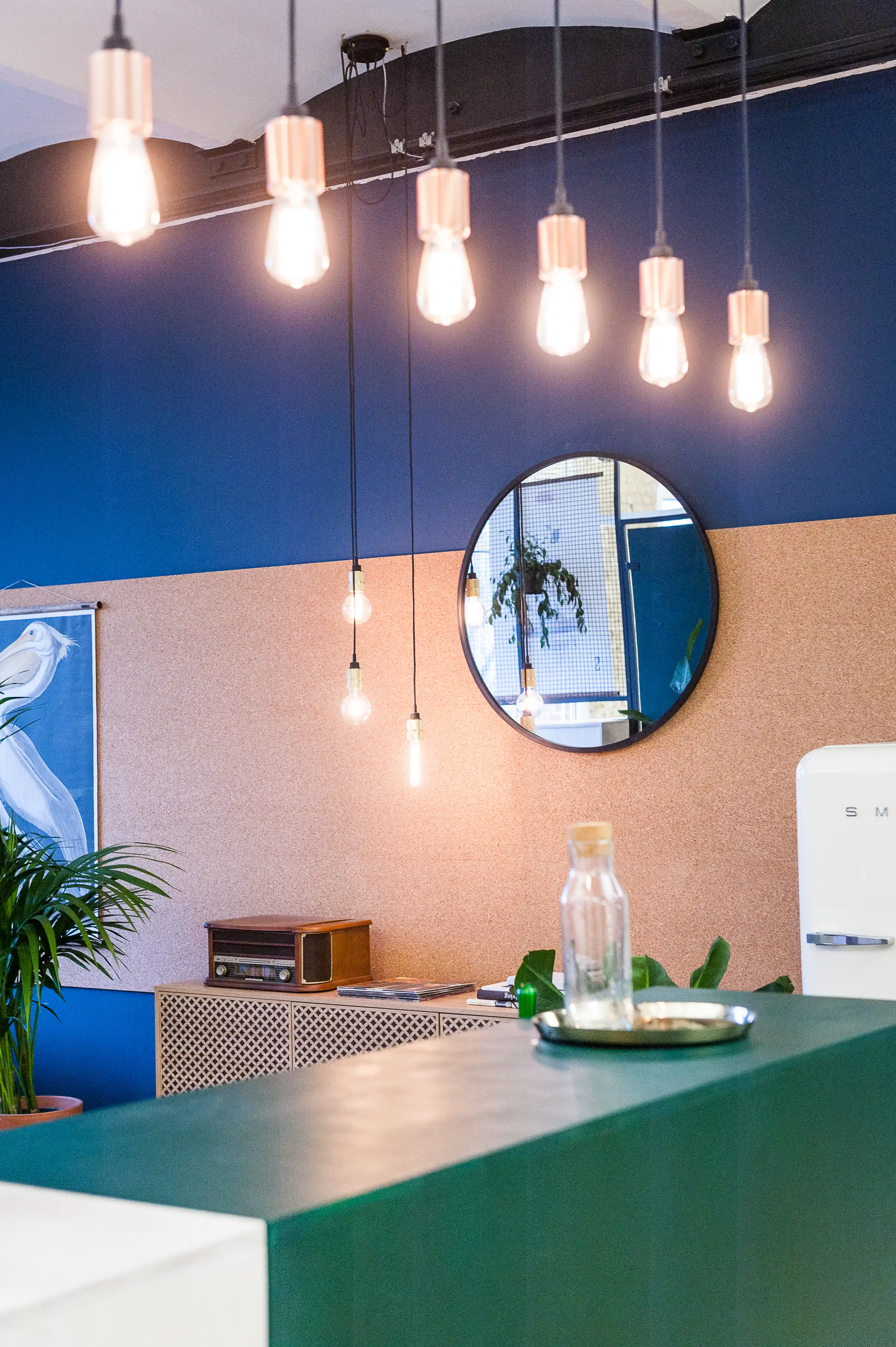 Detail of the kitchen counter at INTERMATE x TRUEMATES with copper-socket filament bulbs hanging in front of a cork wall and dark blue paintwork, a round mirror, vintage radio and white retro fridge.