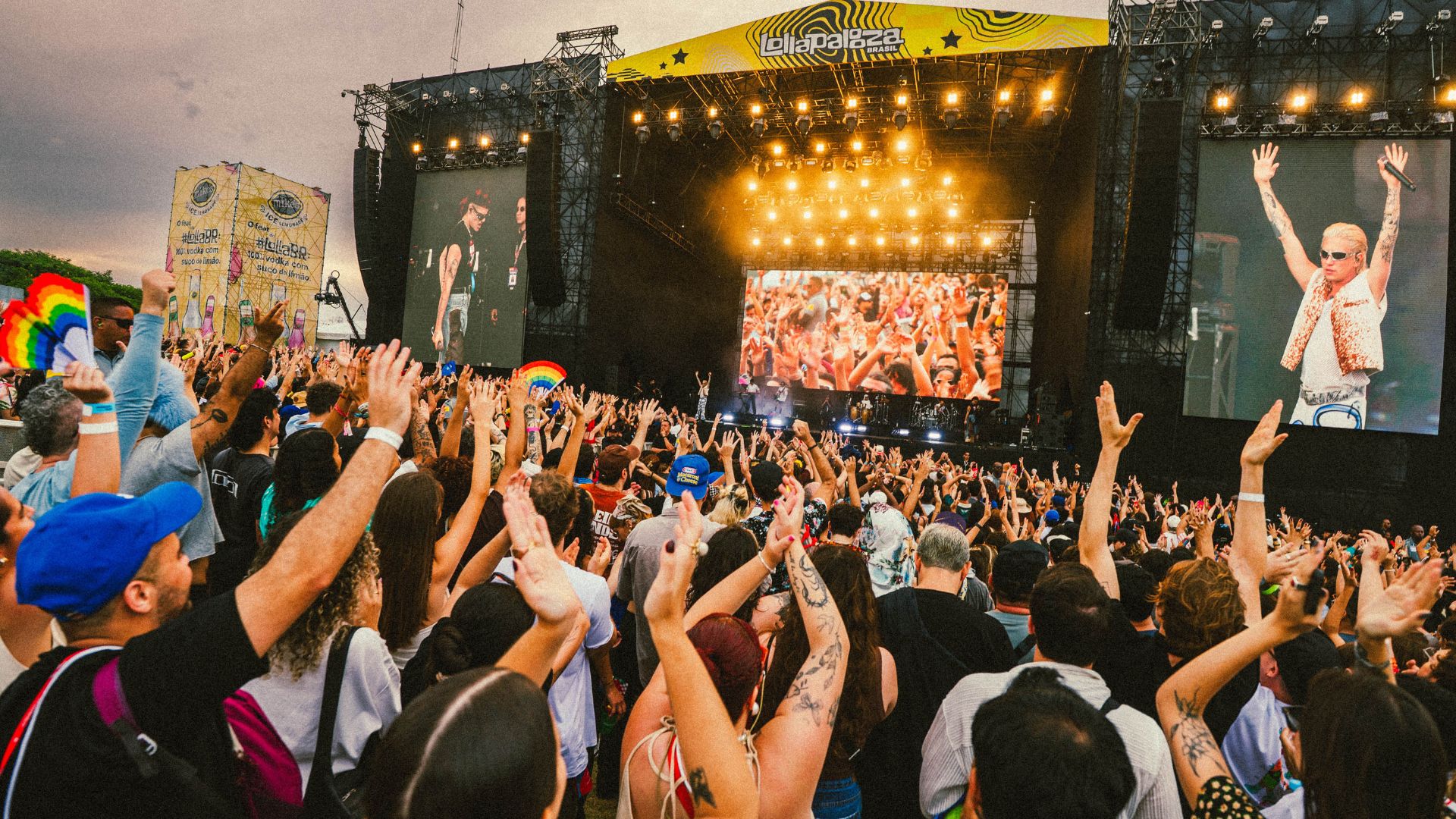 Multidão com braços levantados assistindo a um show ao ar livre, com palco grande, telão e iluminação intensa.