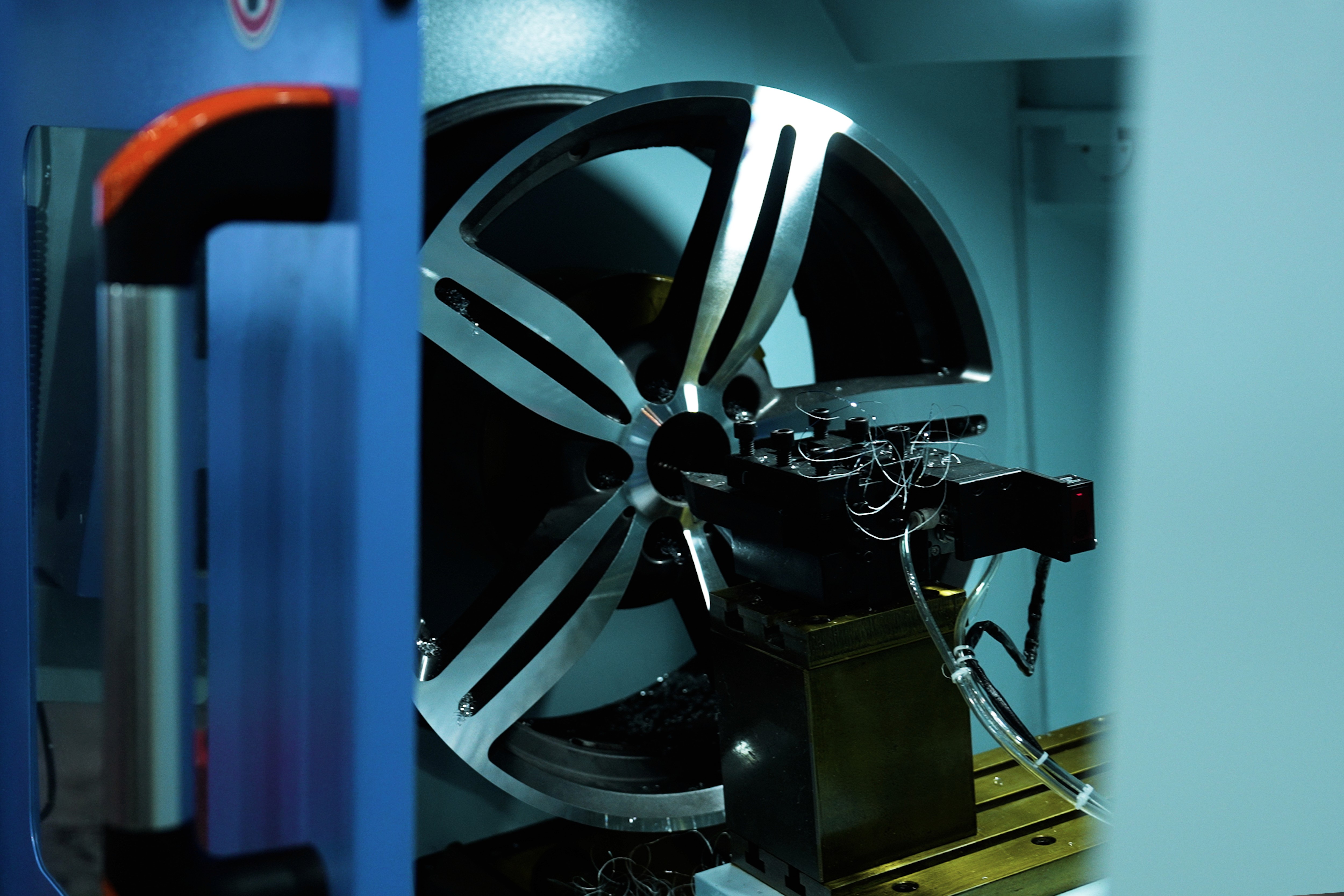 A modern hallway features vibrant teal and orange geometric and leaf patterns, with glossy tiled floors, a large rectangular light fixture, and a small shelf holding a potted plant, all leading to a large, frosted window at the end.