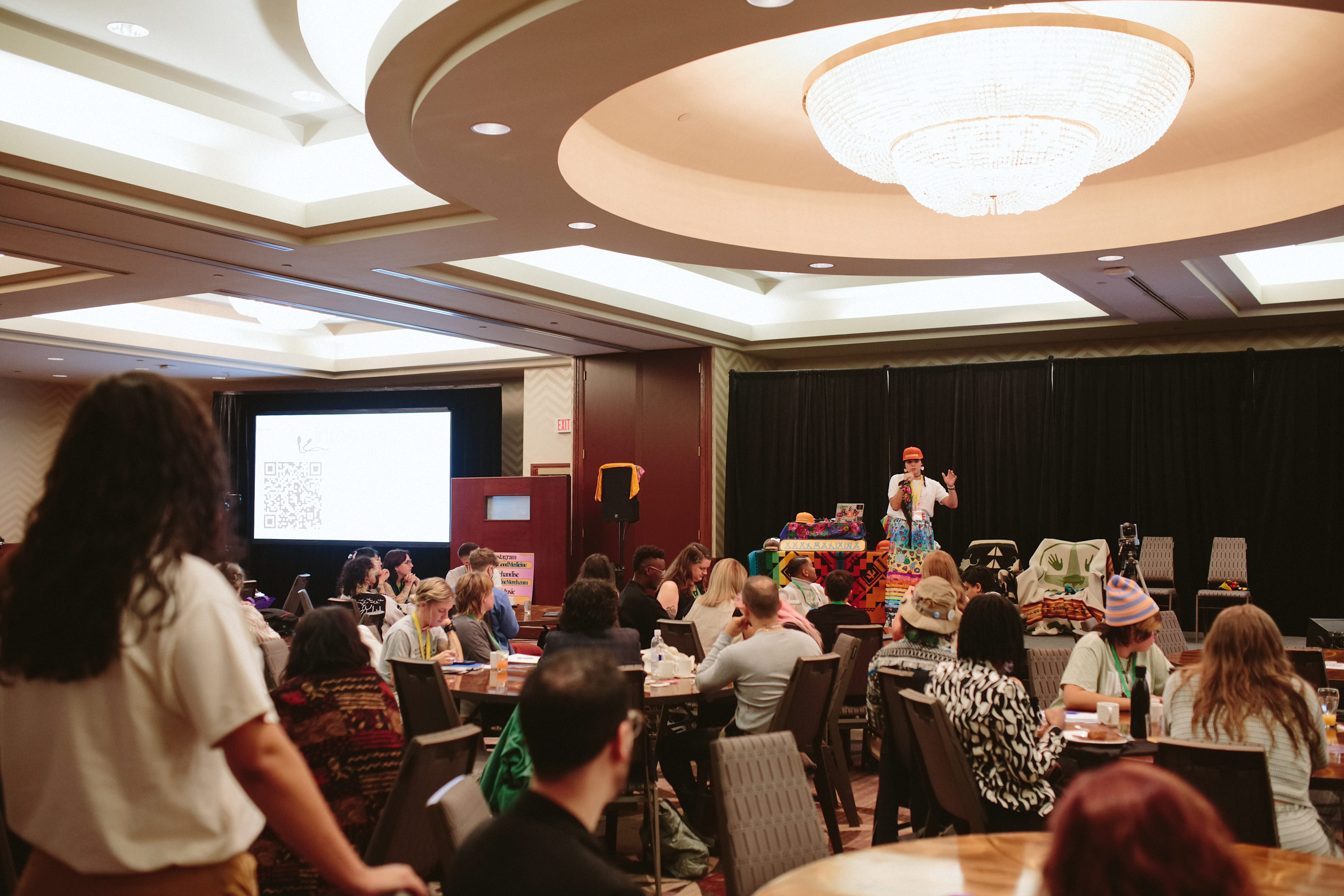 Un groupe diversifié de personnes assises à des tables rondes, l'arrière d'un individu debout tourné vers la scène. Tous regardent une personne en jupe colorée prendre la parole.