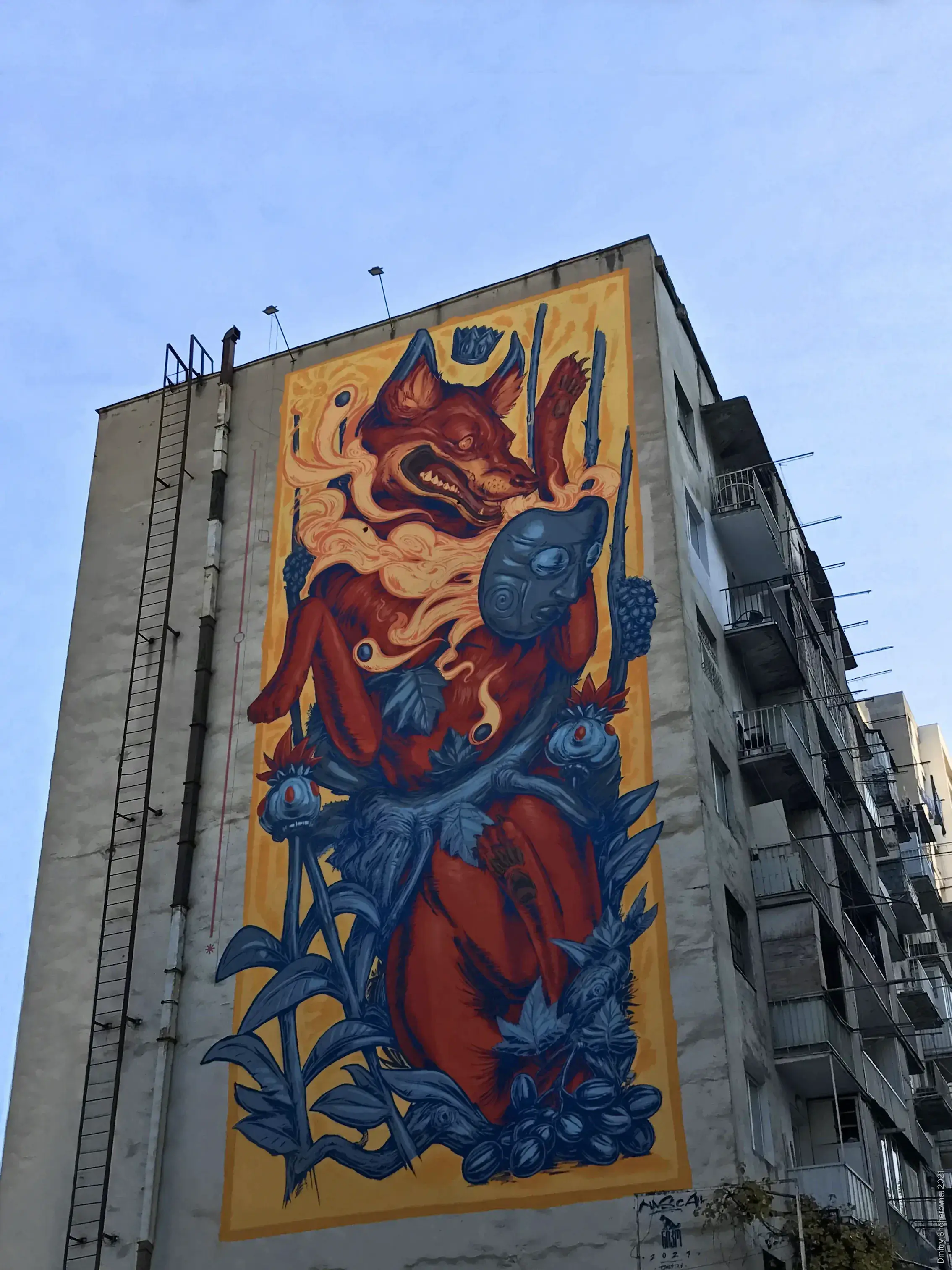 A multi-story building with a mural depicting a snarling red fox holding a blue tribal mask, surrounded by blue foliage and wide-eyed fish, set against a golden yellow background.