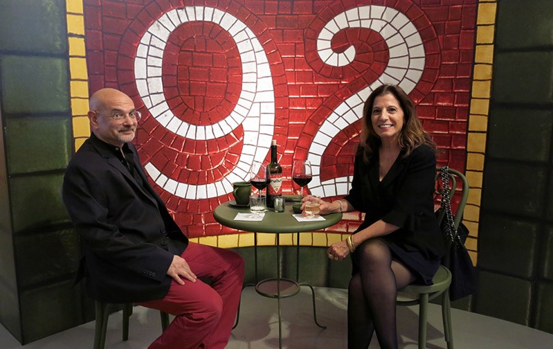 picture of a men and woman sitting at a table and behind them is a red wall with no. 92