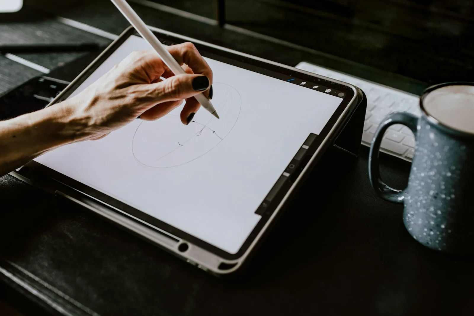 Hand with dark nail polish sketching a circle with a stylus on a tablet, next to a speckled mug and keyboard