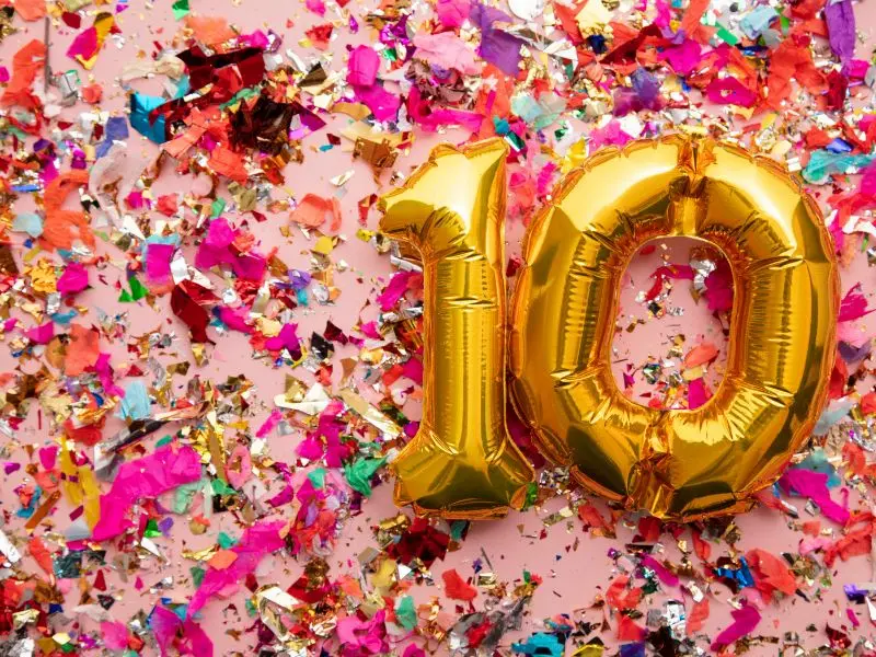 Golden balloons in the shape of the number 10 are set against a backdrop of scattered multicolored confetti pieces on a soft pink surface, symbolizing a vibrant celebration of a 10-year milestone.