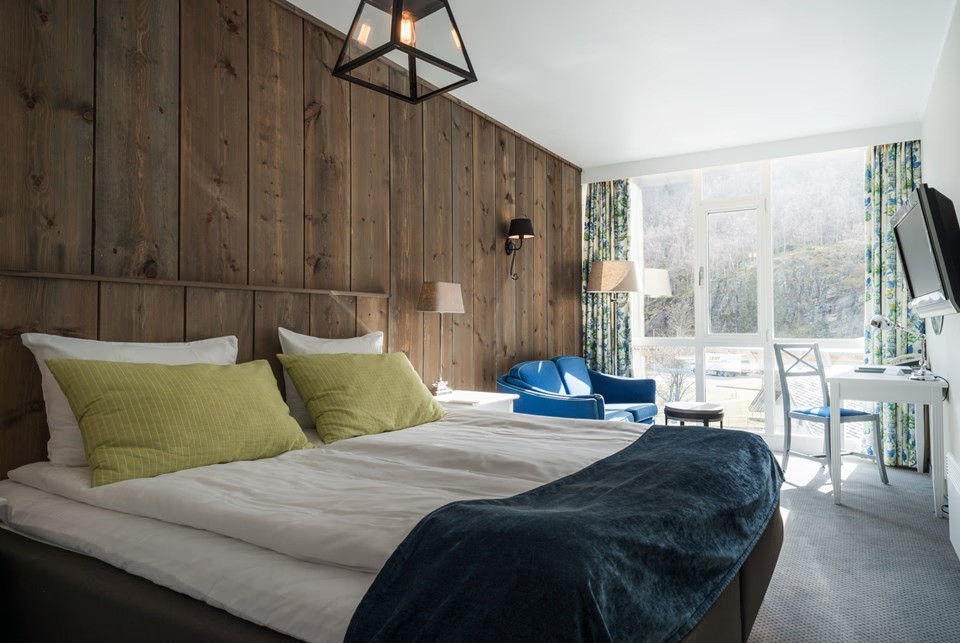 A beautifully designed hotel room features a large double bed with a patterned blue headboard, adorned with decorative pillows, set against a floral accent wall, and complemented by a window view of lush greenery, alongside two plush lounge chairs and a circular table.