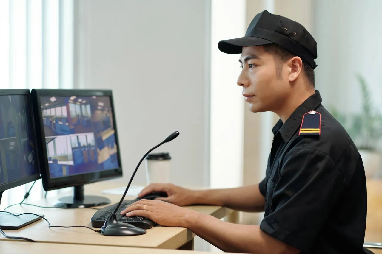 A security guard in uniform sitting at a desk watching security feeds on a monitor.
