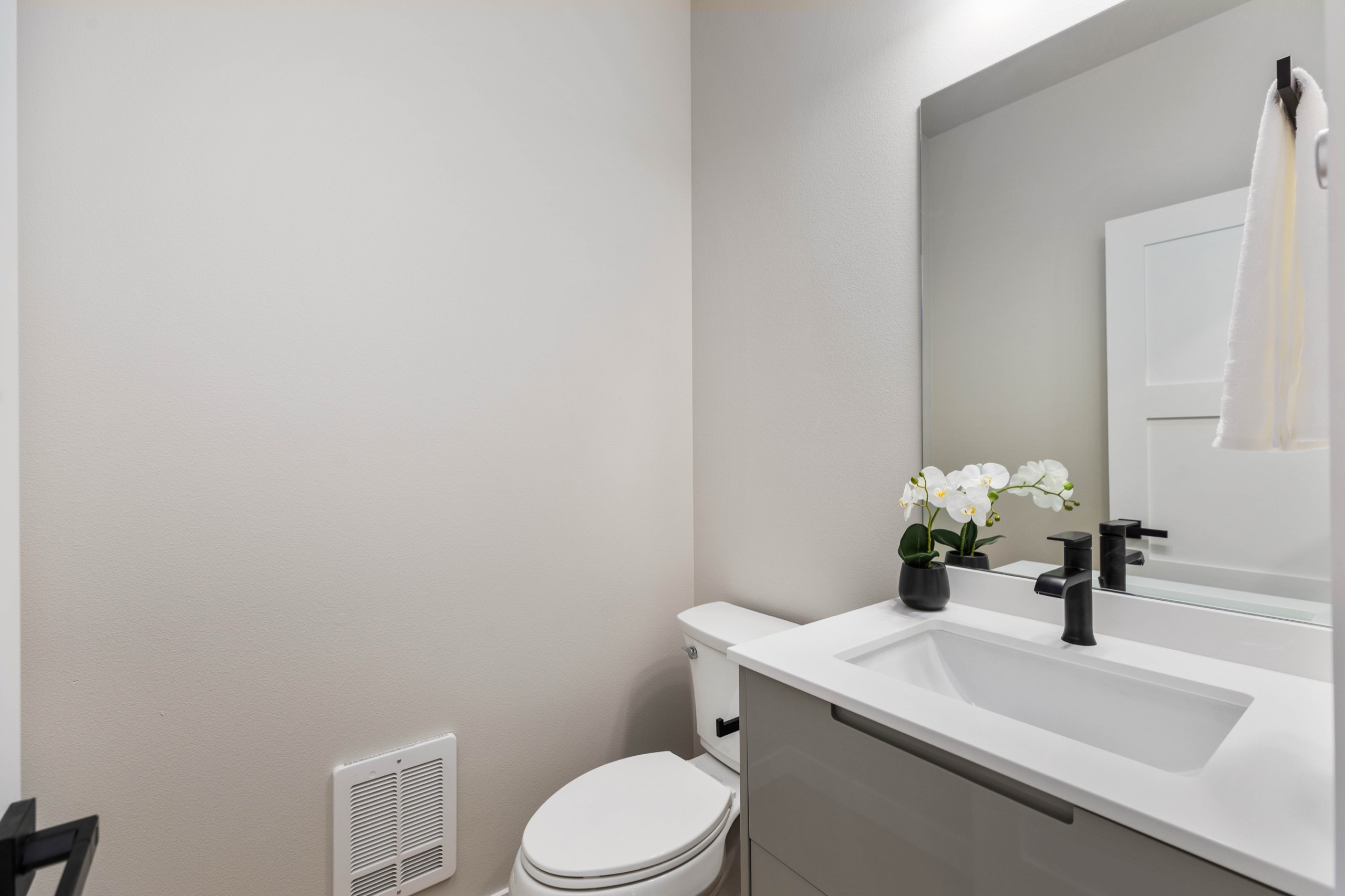 Clean and modern luxury half bathroom with grey vanity, white countertop, black fixtures, oversized frameless mirror, and white orchid floral accent