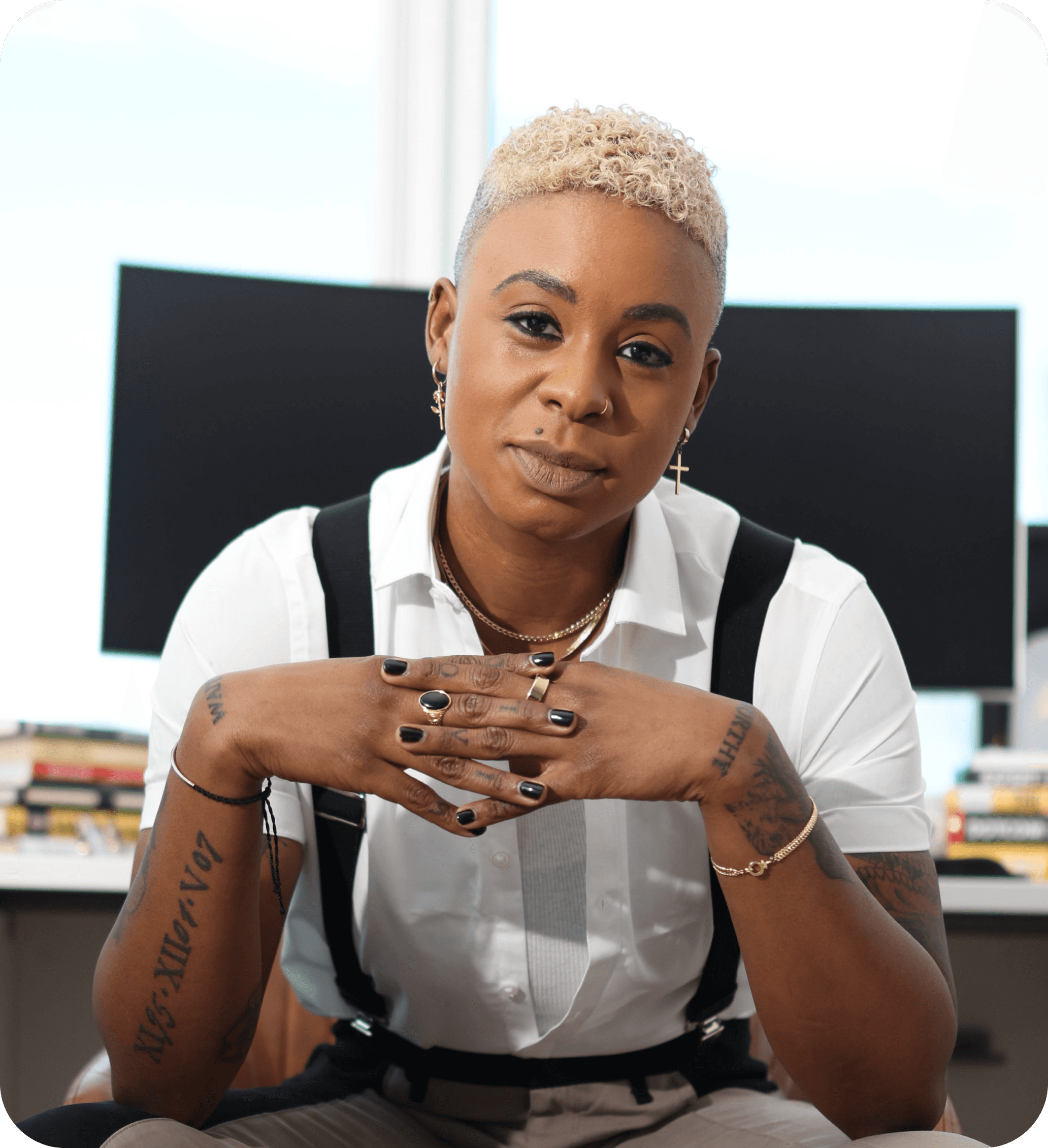 Ashleigh Warren, a person with short blonde hair sits at a desk, looking confidently at the camera, hands clasped together.