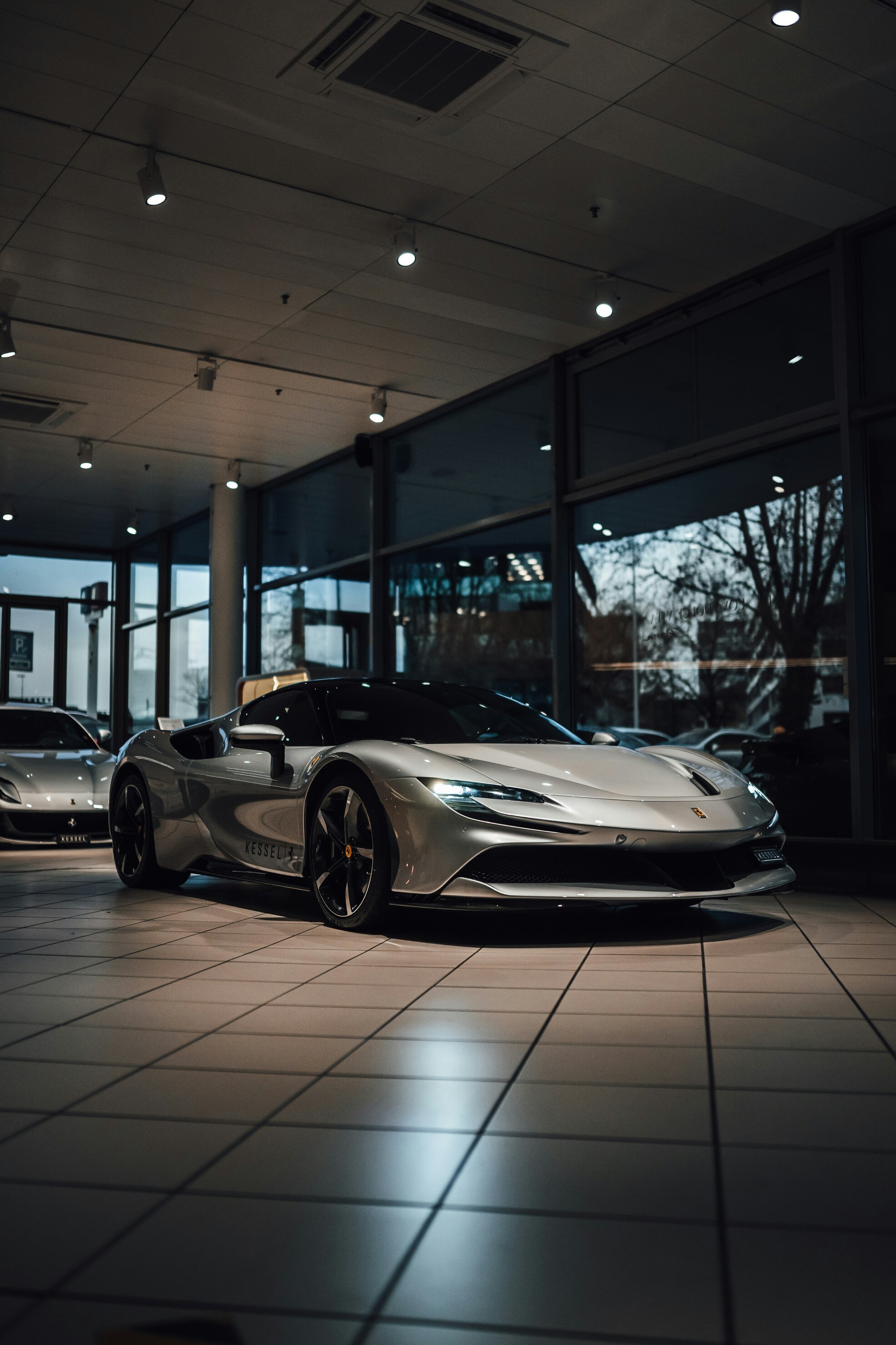 A sleek, silver sports car is displayed in a showroom with floor-to-ceiling windows, reflecting modern architecture and trees outside.