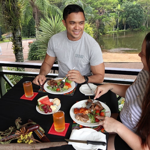 Two people enjoy a meal at an outdoor table with plates of food, drinks, and fruit. Lush greenery and a pond are visible in the background.