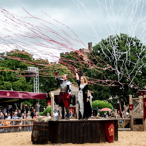 Kaksi esiintyjää keskiaikaisissa puvuissa seisoo puisella korokkeella ja vilkuttavat väkijoukolle. Punaiset ja valkoiset nauhat täyttävät taivaan.