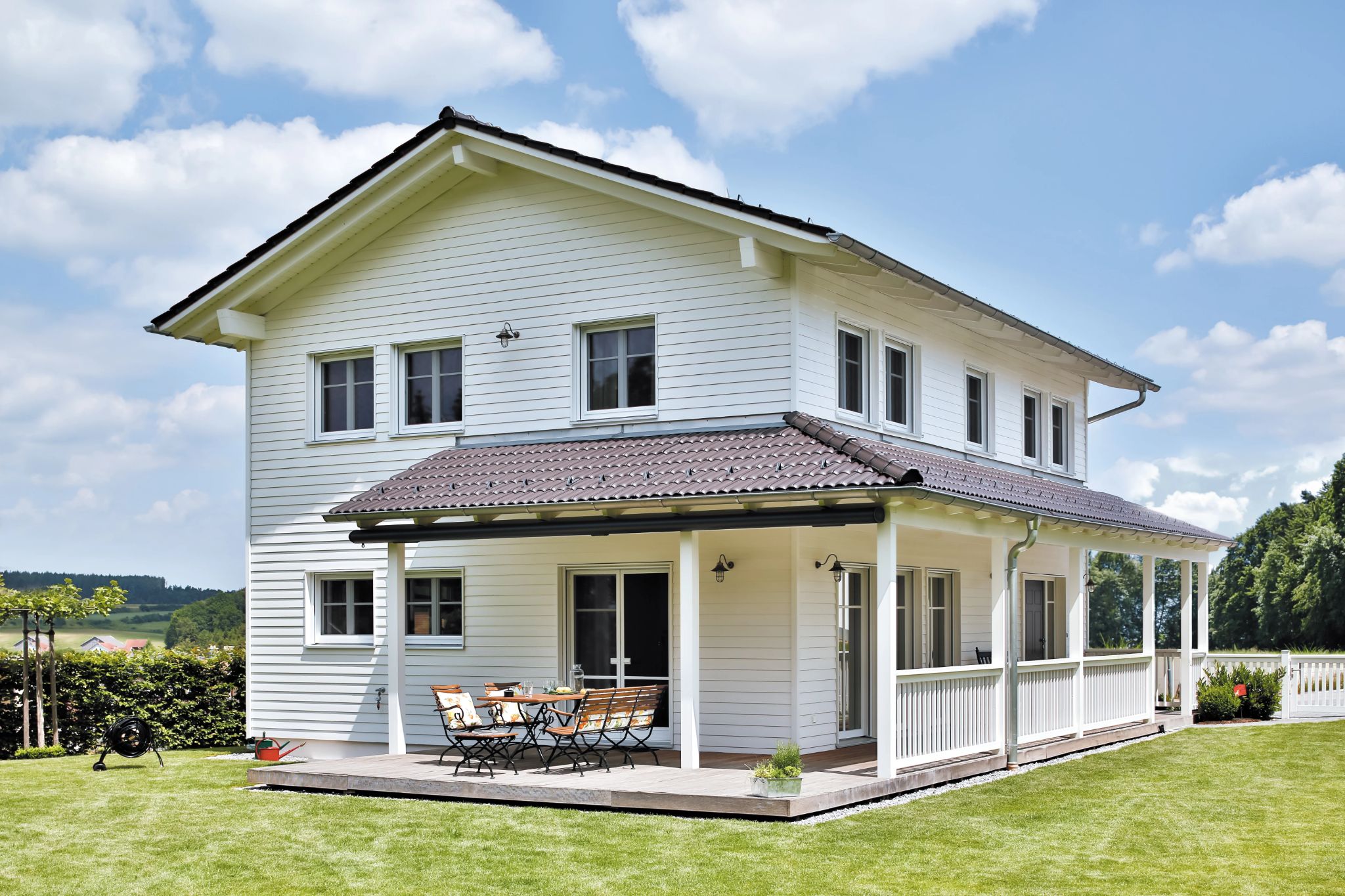 Modernes weißes Holzhaus mit überdachter Terrasse, Sitzgruppe und großem, gepflegtem Rasen.