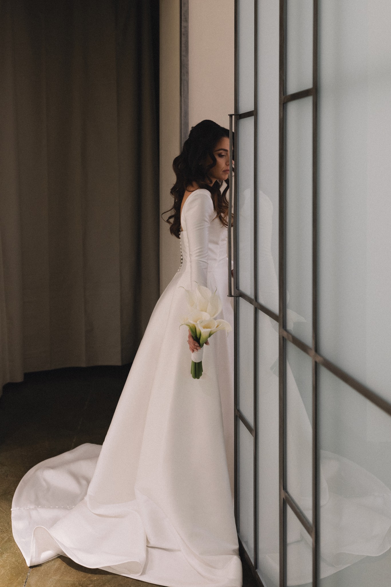 Sposa in mikado bianco con bouquet di calle, profilo davanti a vetrate geometriche, preparativi matrimonio a Parma