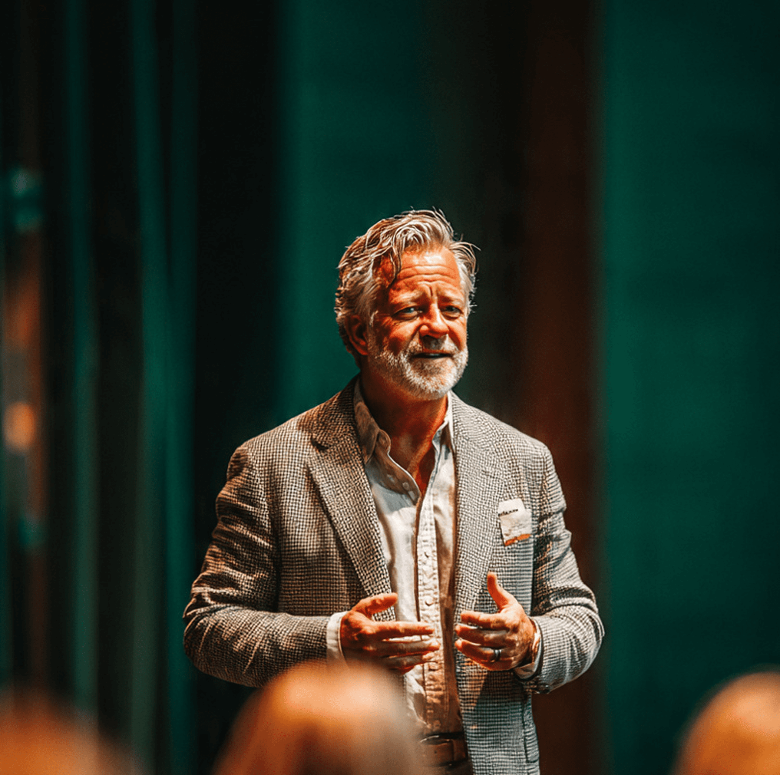 Older man with gray hair and beard in a tweed blazer speaking and gesturing on a stage with a green backdrop.