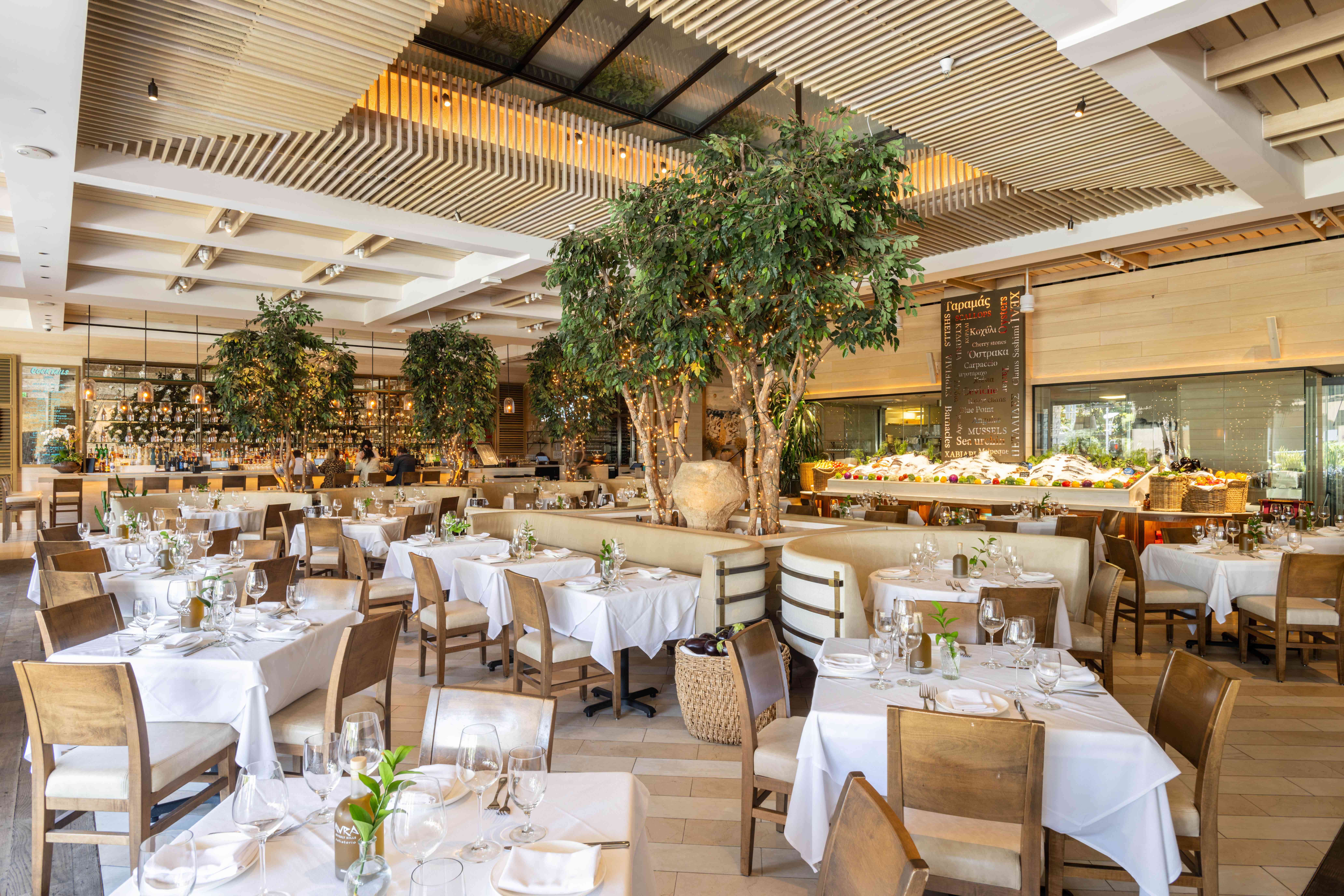 A spacious restaurant interior featuring white tablecloths, wooden furniture, and green plants.