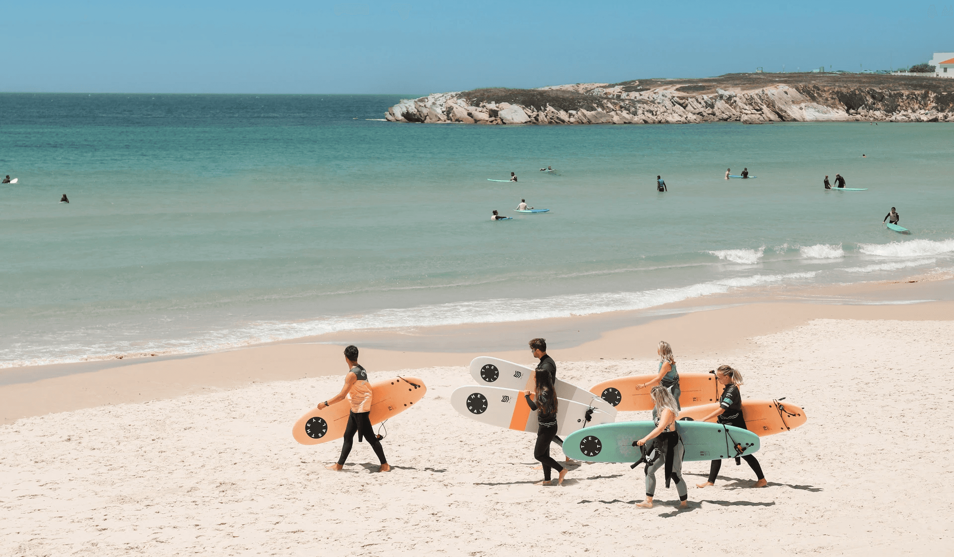Surf students walking on Peniche beach carrying surfboards branded with The Salty logo.