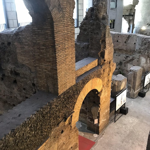 Rovine di antichi mattoni con arcate e pareti parzialmente in piedi, situate in un ambiente interno simile a un museo.