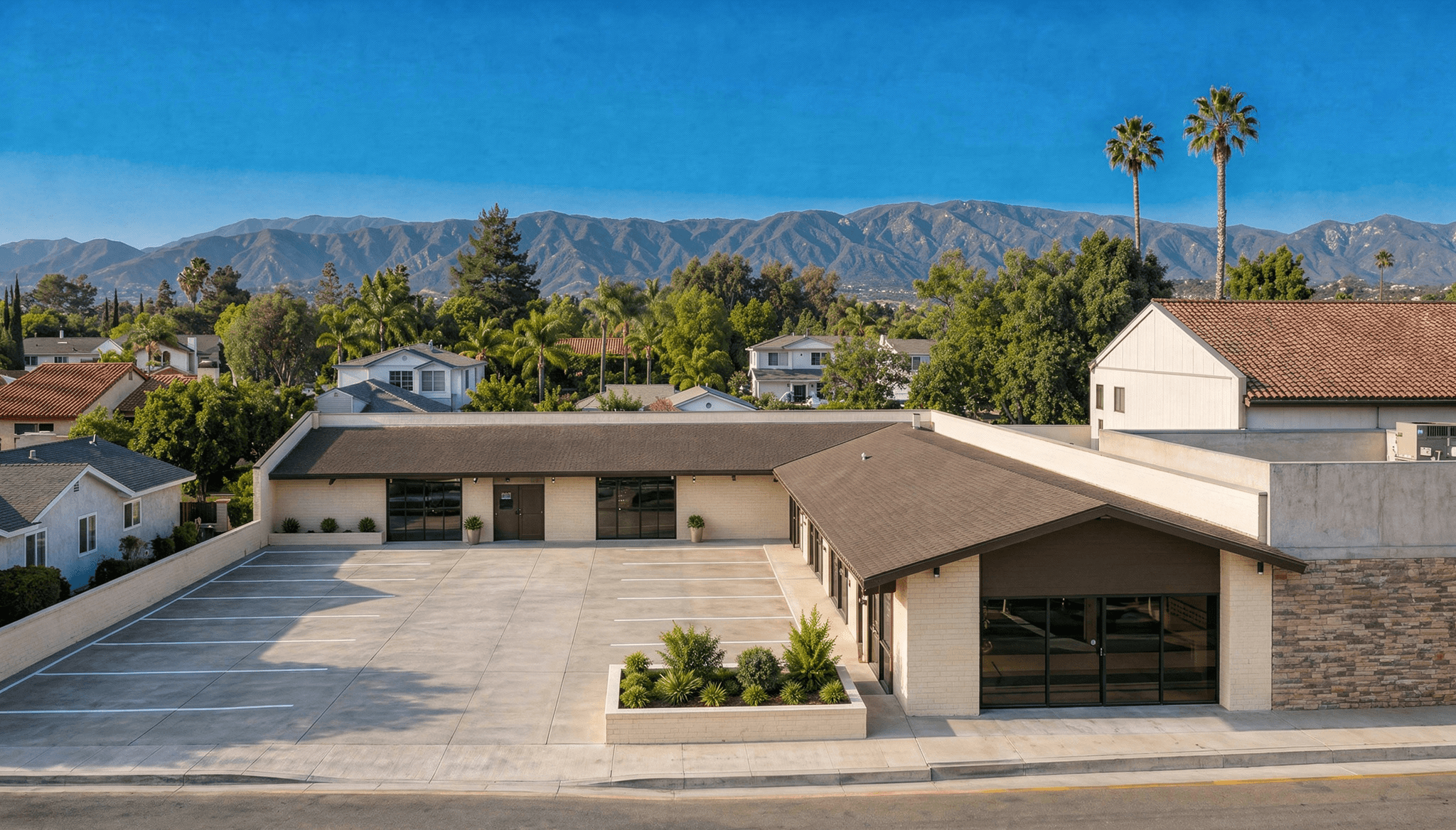 Street-level exterior of a renovated single-story commercial building with a low-pitched gabled roofline, warm brown standing-seam metal roof accents, stone veneer facade panels, large glass storefront entry, a wide paved parking apron with landscaped planters, and a backdrop of tall palm trees and snow-dusted mountain peaks under a clear blue sky.