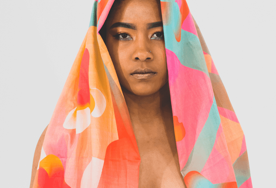 A person with dark skin and dark hair holds a brightly colored, patterned scarf over their head and shoulders. The scarf features abstract shapes in orange, pink, white, and teal against a light gray background. The person’s face is visible, with dark eyes and dark eyebrows, and their expression is neutral. The background is a plain light gray.