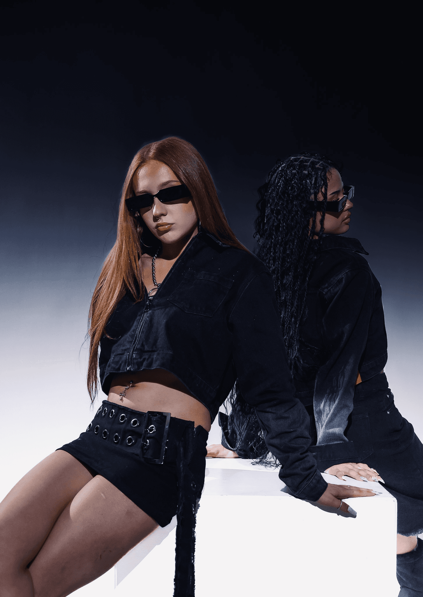 Two women dressed in edgy black outfits and rectangular sunglasses pose back-to-back against a gradient background