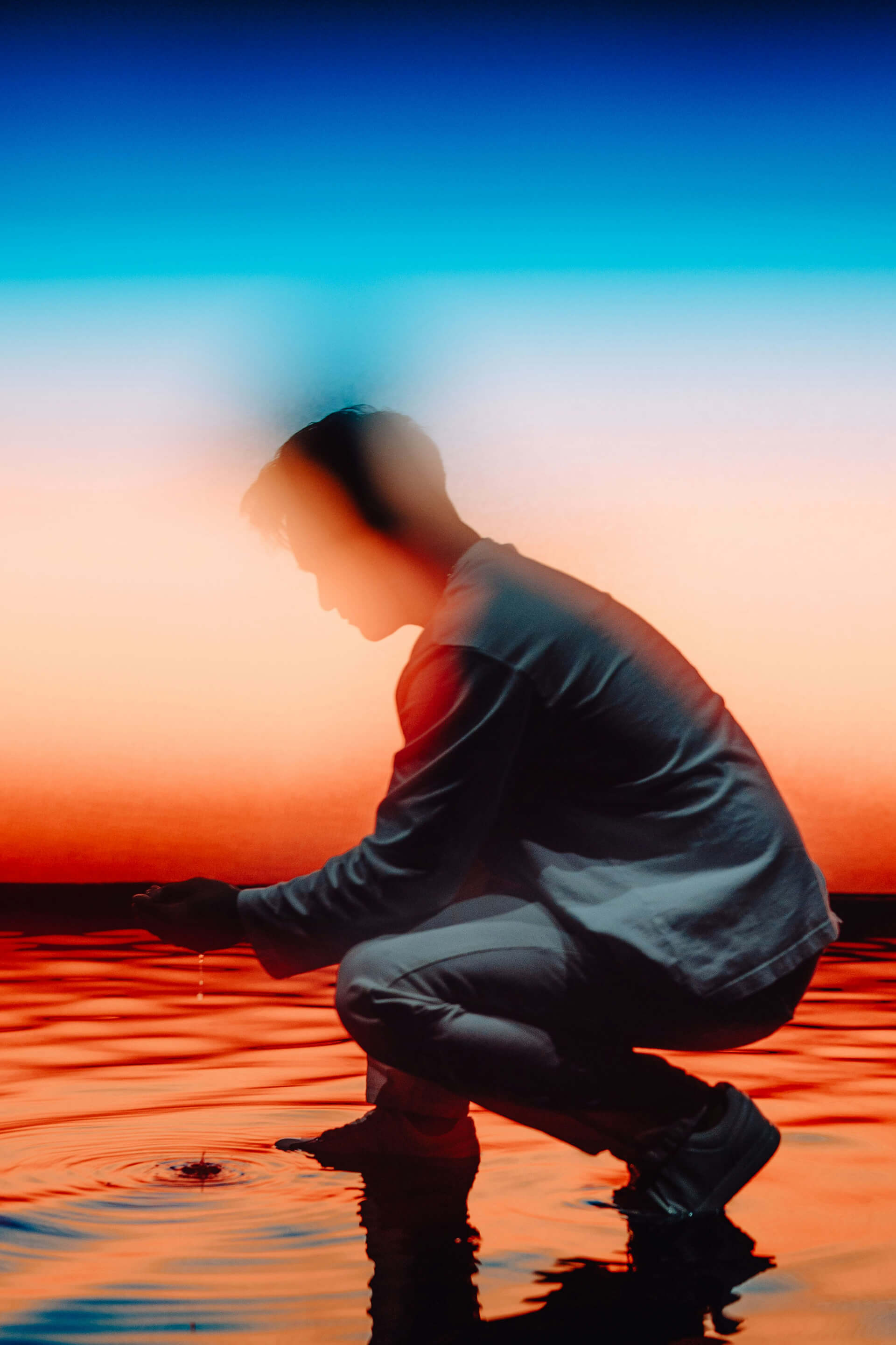 A person in white clothing crouches at the edge of a body of water, cupping their hands as droplets fall and create ripples. The background glows in a surreal gradient of deep blue, turquoise, and vivid orange, casting an ethereal and dreamlike atmosphere.