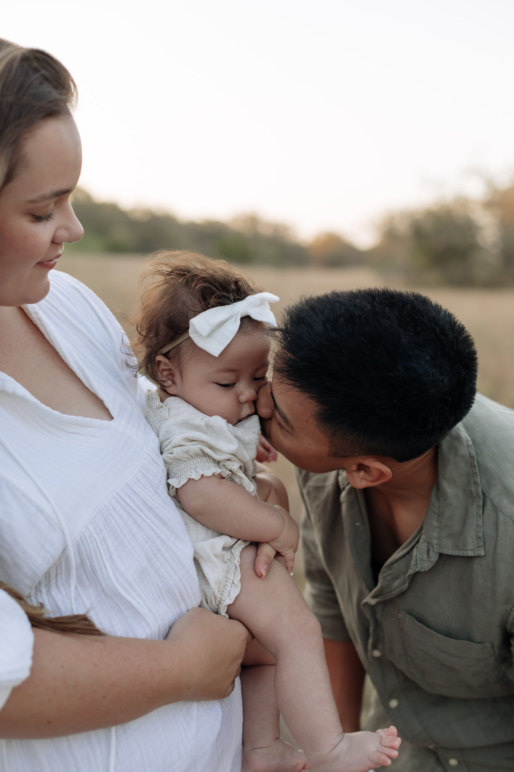 Candid Mackay family session at golden hour with dad kissing baby girl in an open grassy landscape