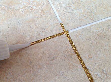 A close-up of a person applying gold glitter tile grout to a beige ceramic tile joint. The grout adds a decorative, sparkling effect to the tile design.