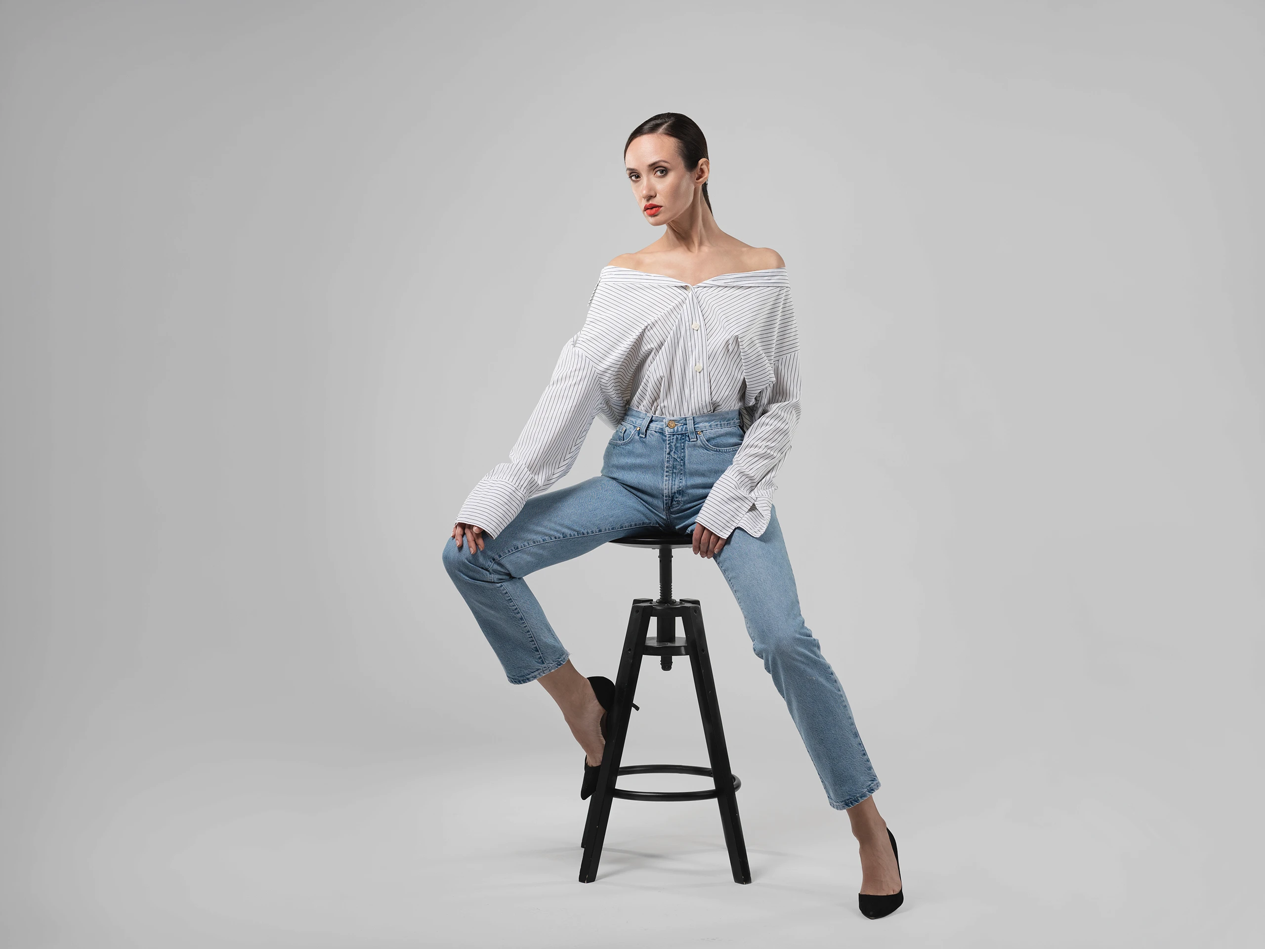 Minimal fashion portrait of a woman seated on a stool, wearing an off-shoulder blouse and jeans.