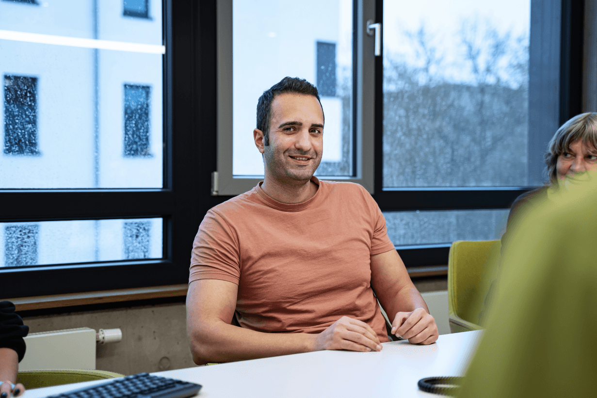Mann in rosafarbenem T-Shirt sitzt lächelnd in einem modernen Besprechungsraum der kba Architekten und Ingenieure GmbH; große Fenster mit Regen im Hintergrund und weitere Personen am Tisch.
