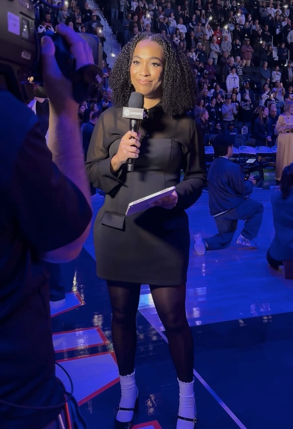Autumn Johnson in a black dress holds a microphone and notepad while standing on a basketball court, facing a cameraman. The background shows a crowd seated in the arena, with blue lighting illuminating the scene.