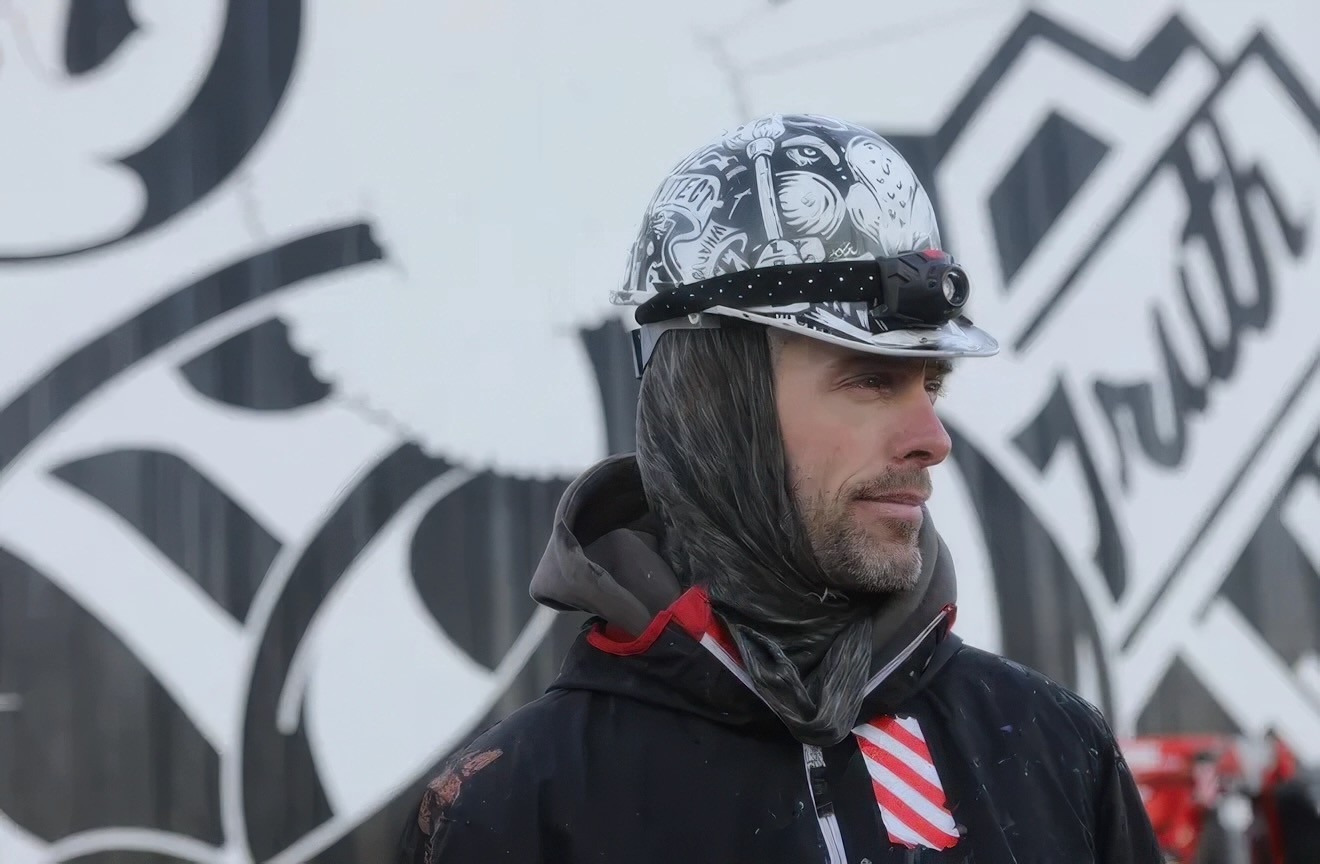 A man in a painted helmet and hooded scarf looks past the camera, a black-and-white lettered wall behind him.